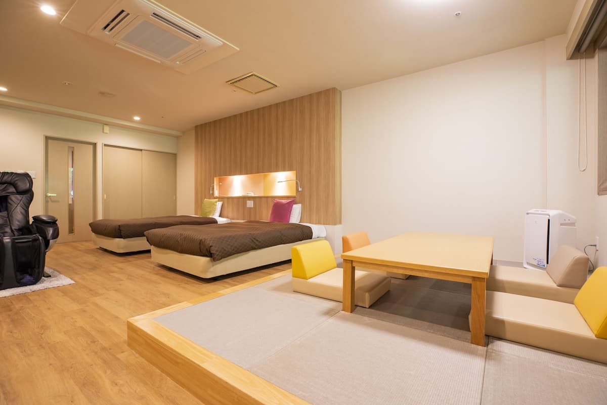 A modern hotel room with a bed, sofa, and a small dining table. There is a desk lamp, a heater, and a suitcase on the floor.