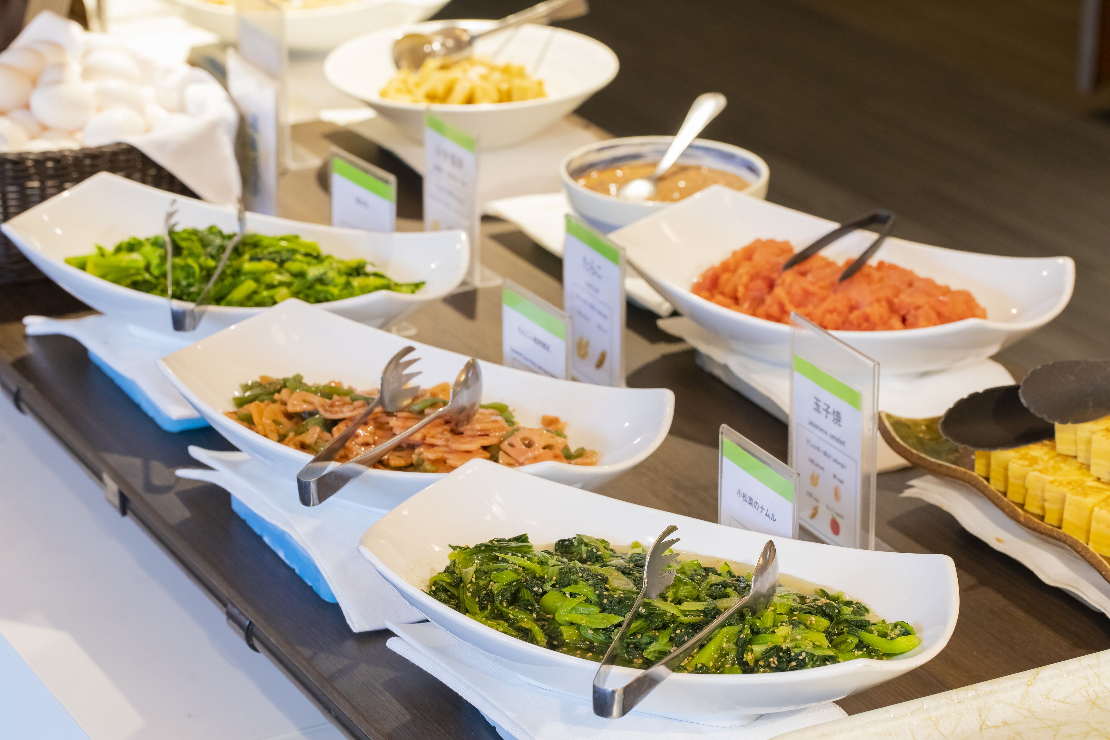 A buffet with various dishes in white bowls, each labeled with a green sign.