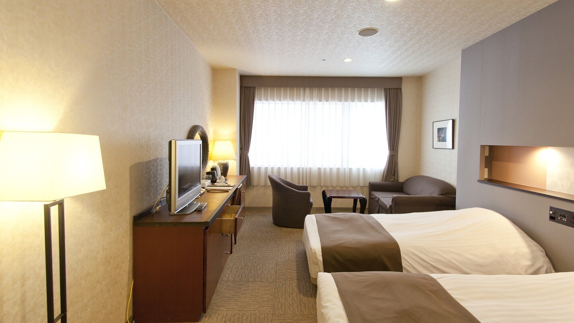 A hotel room with a bed, a desk with a TV, a chair, a sofa, and a window with curtains.