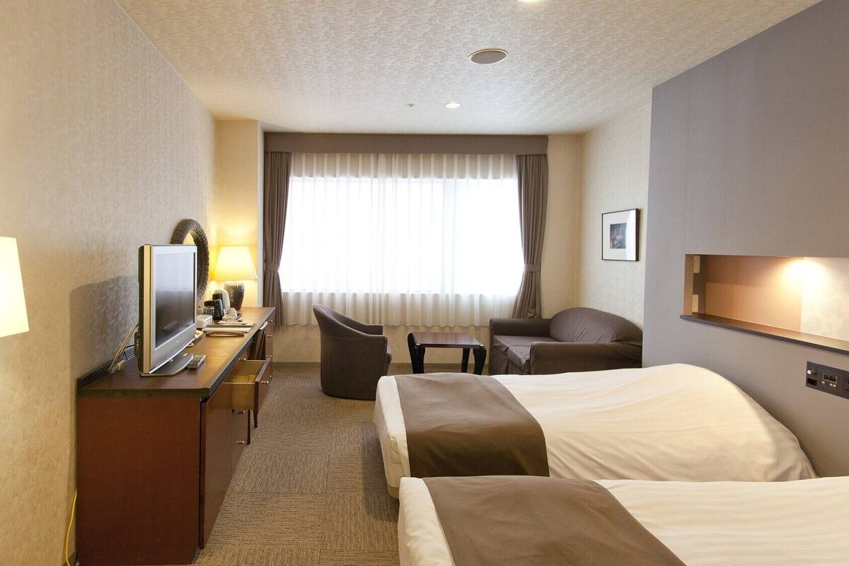 A hotel room with a bed, a desk with a TV, a chair, a sofa, and a window with curtains.