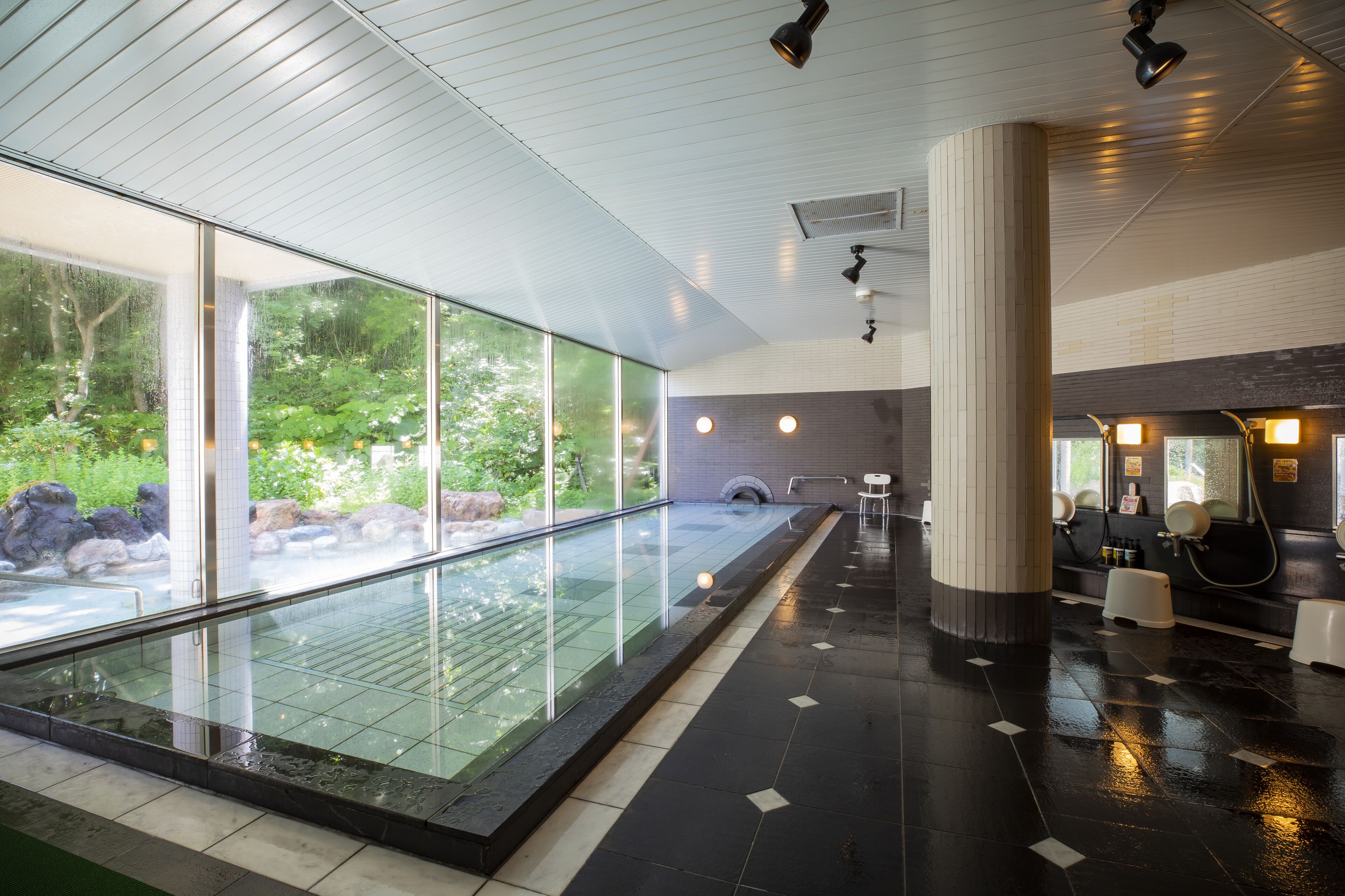 A modern indoor pool with glass walls, a black tiled floor, and a seating area with a view of the outdoors.