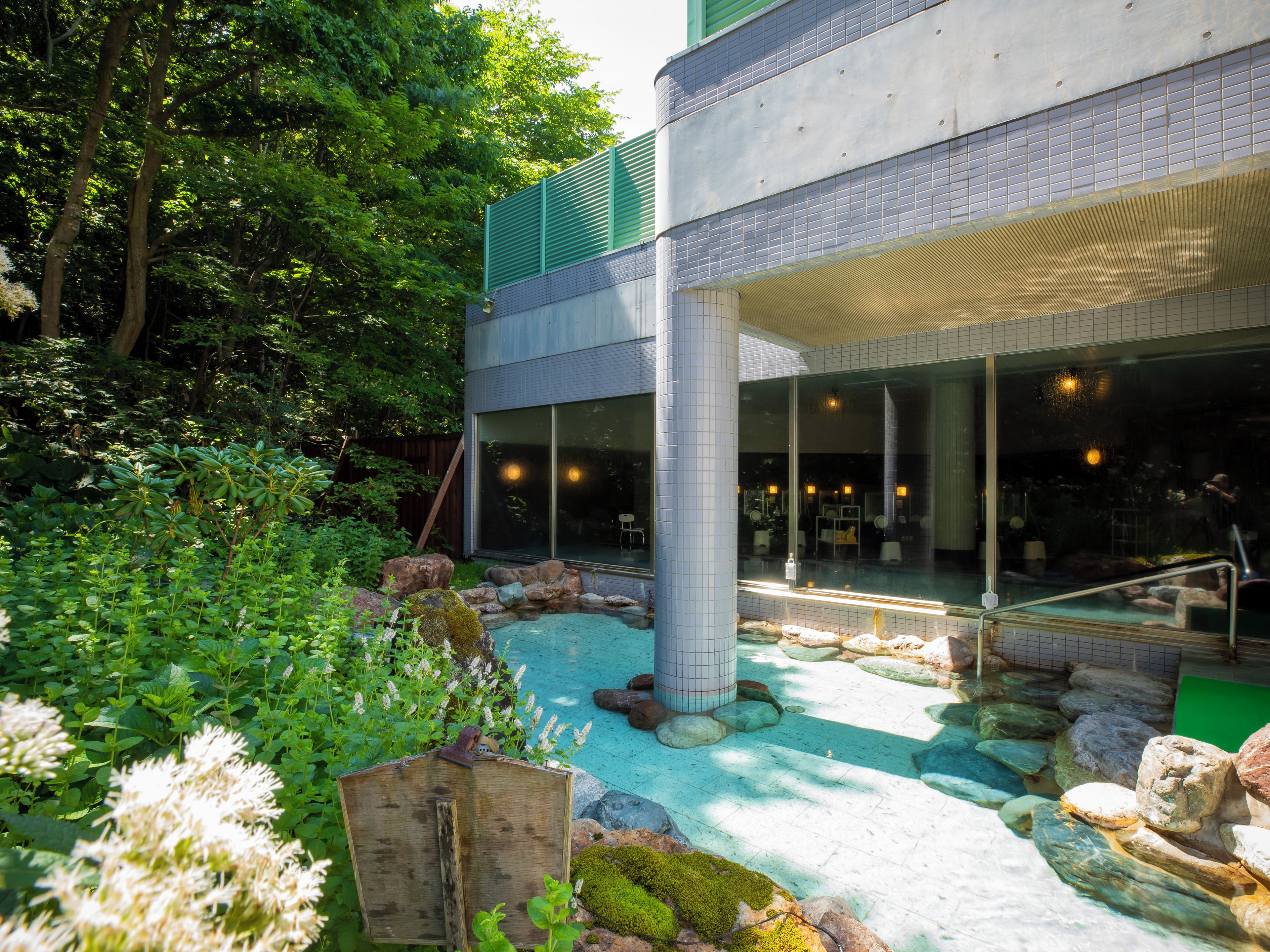 A modern building with a glass facade, a pool area with rocks and plants, and a wooden structure.