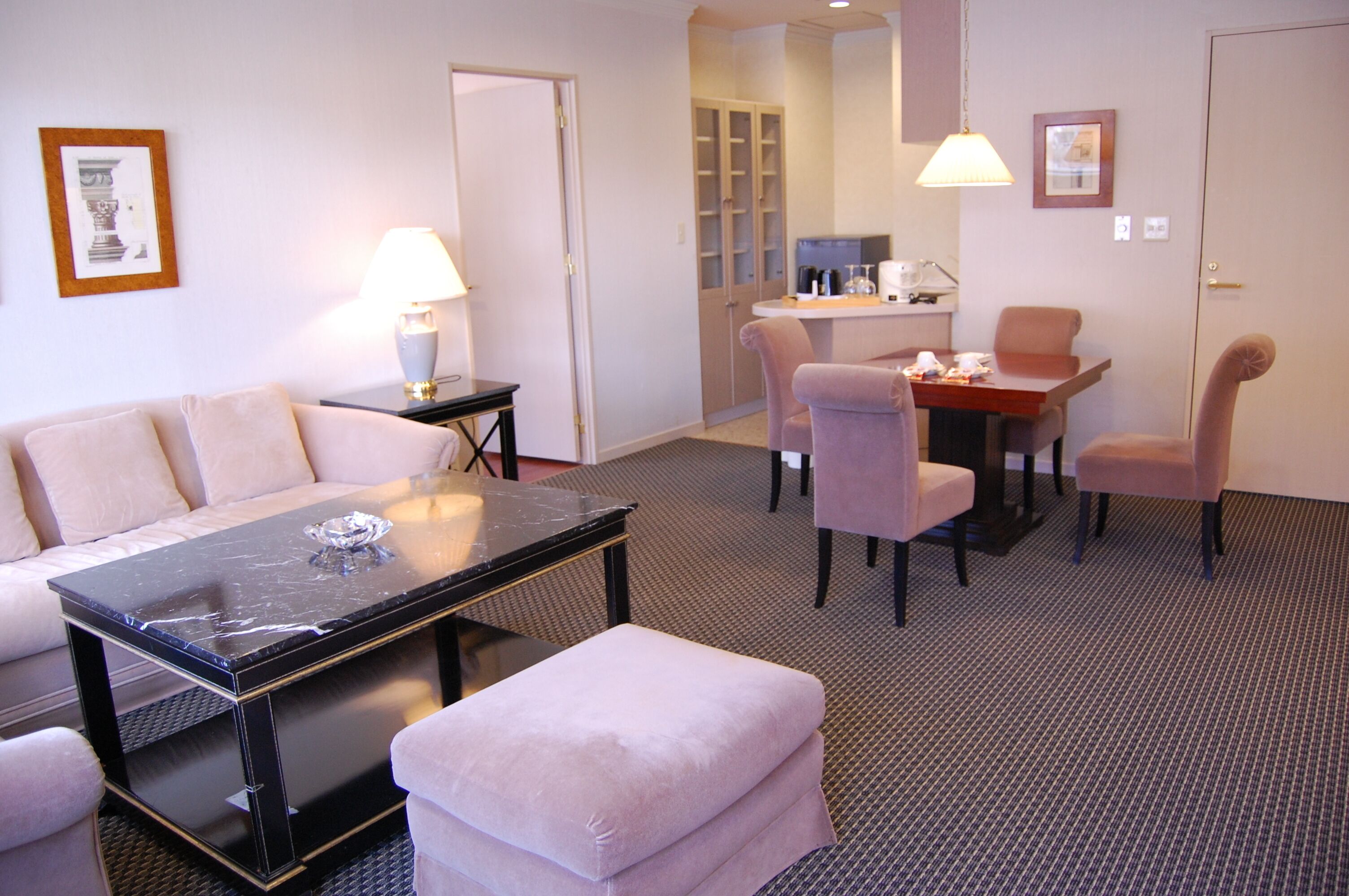 A hotel room with a sofa, a dining table with chairs, a small table with a lamp, and a cabinet.