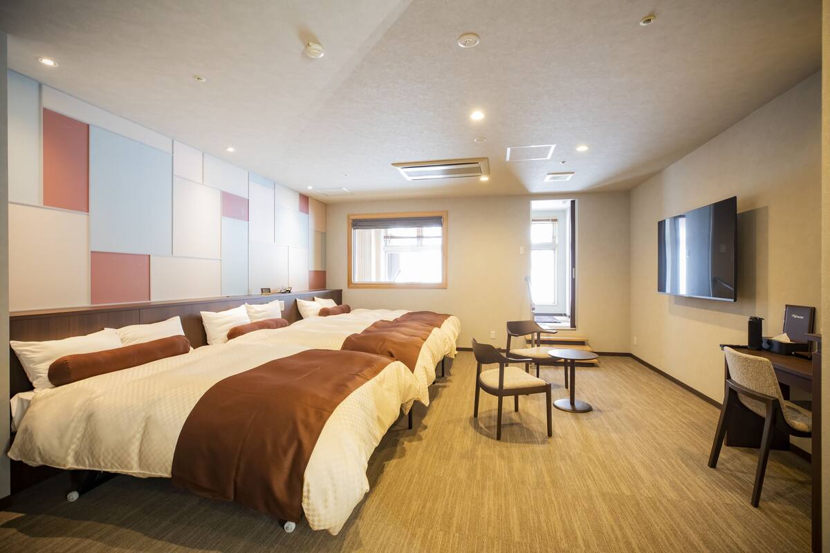 A hotel room with three beds, a desk, a chair, a TV, and a window.
