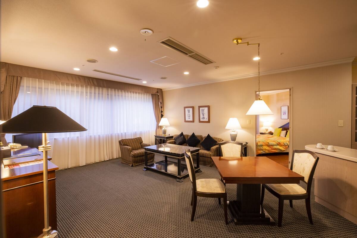 A hotel room with a dining area, a sofa set, a bed, and a desk with a lamp.