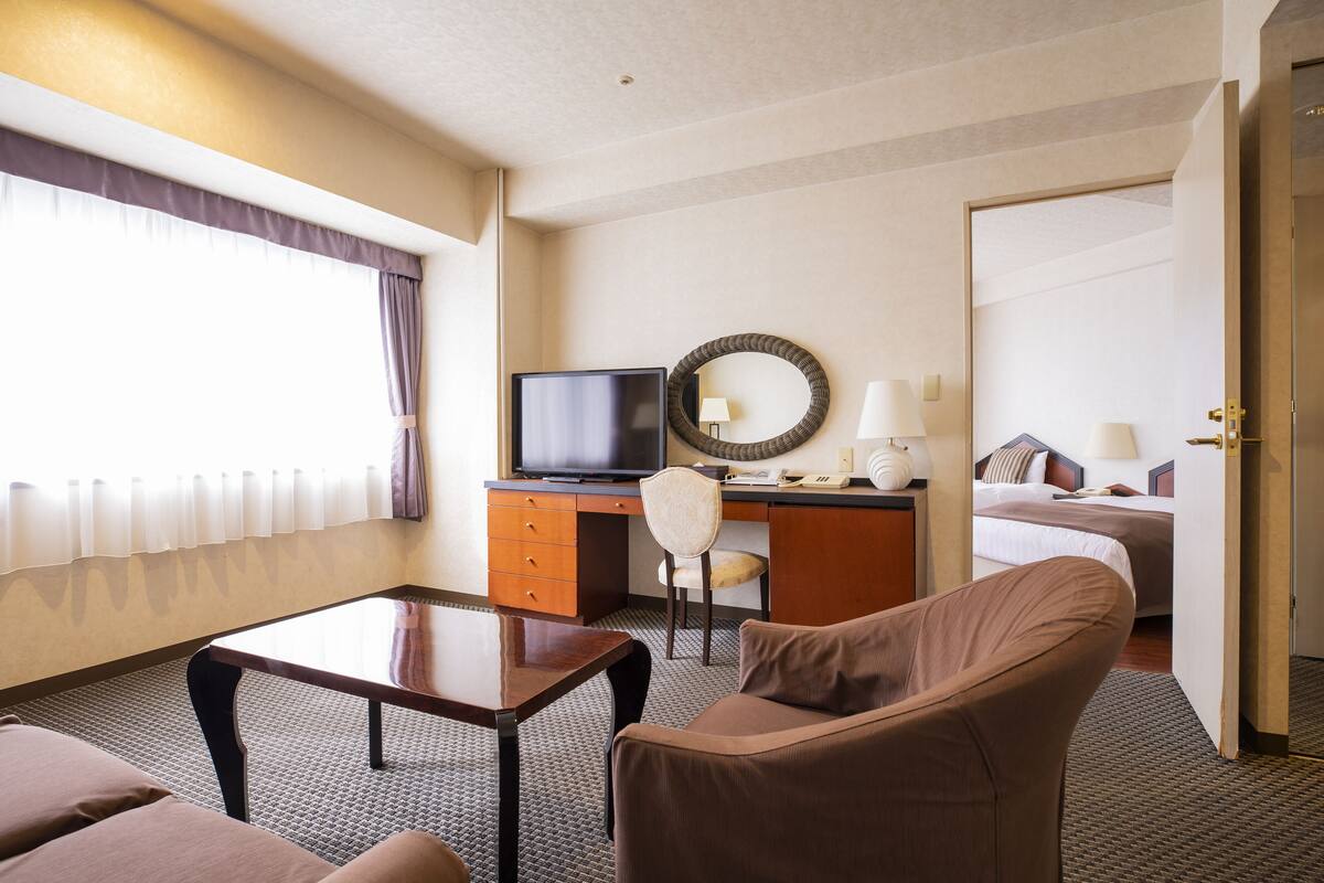 A hotel room with a bed, a desk with a television, a chair, a table, and a lamp.