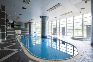 En indendørs swimmingpool med flisegulv, et pool med blå farve og store vinduer der lukker naturligt lys ind.
