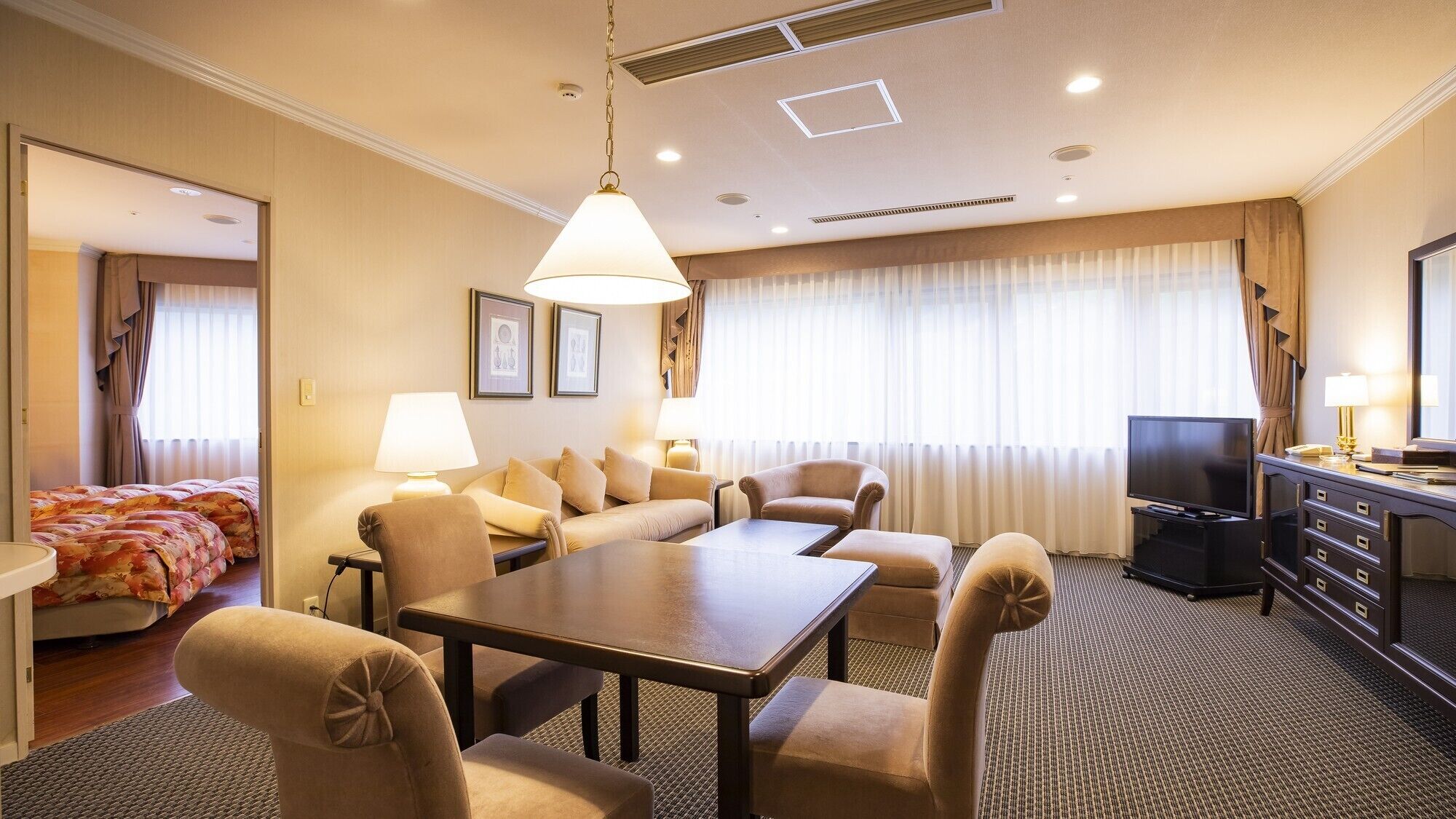 A hotel room with a sofa set, a dining table, a TV, and a desk with drawers.