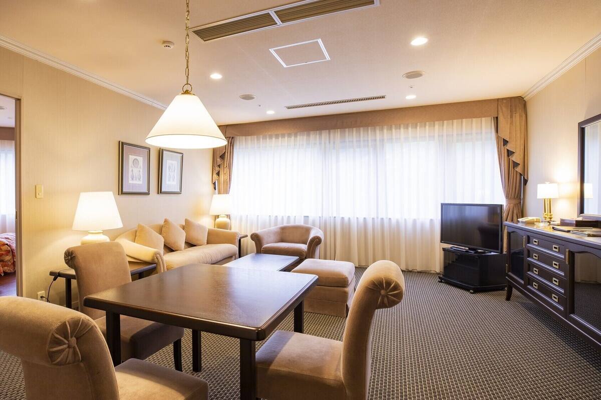 A hotel room with a sofa set, a dining table, a TV, and a desk with drawers.