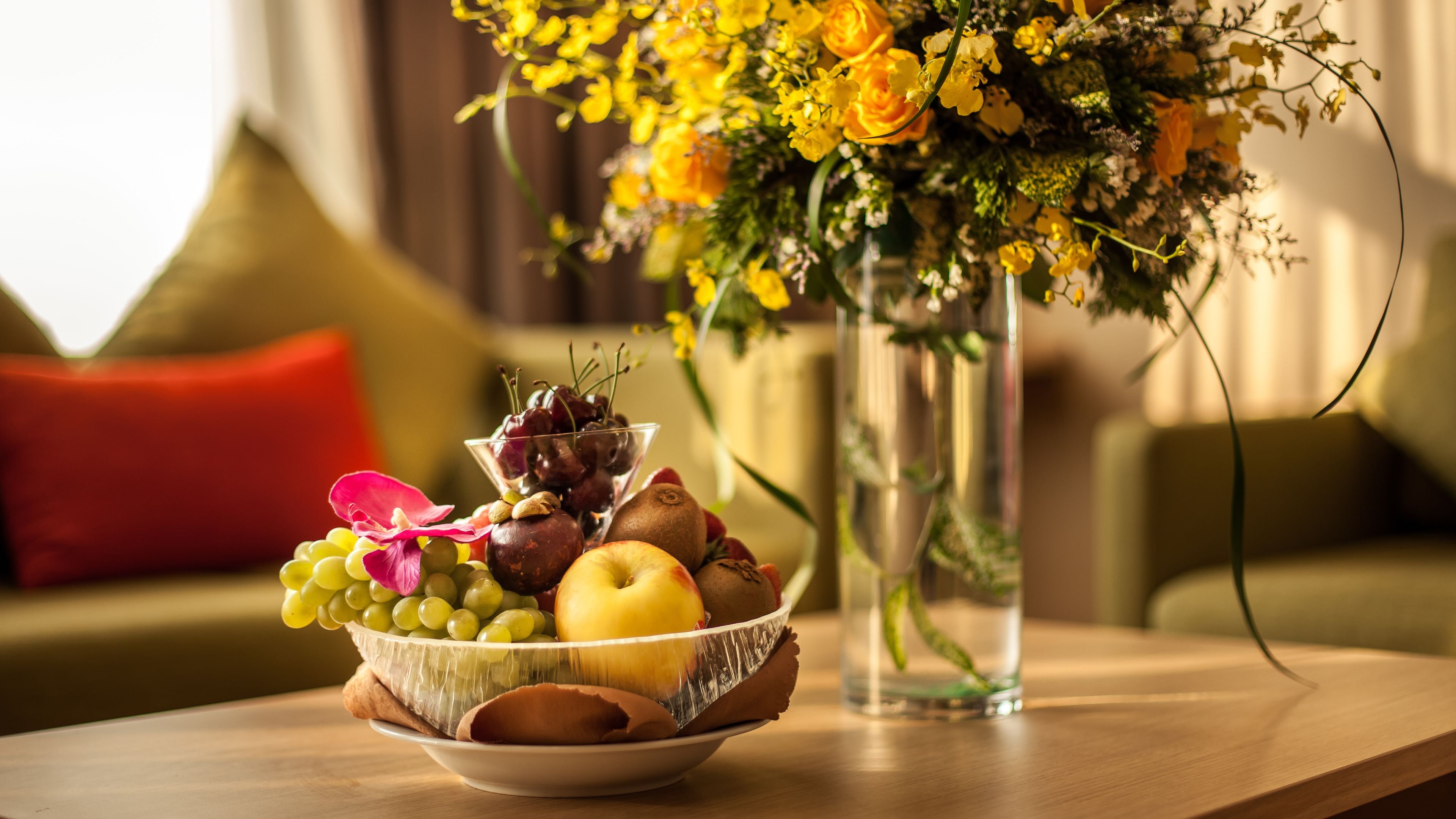 A fruit bowl on a table with a vase of flowers in the background.