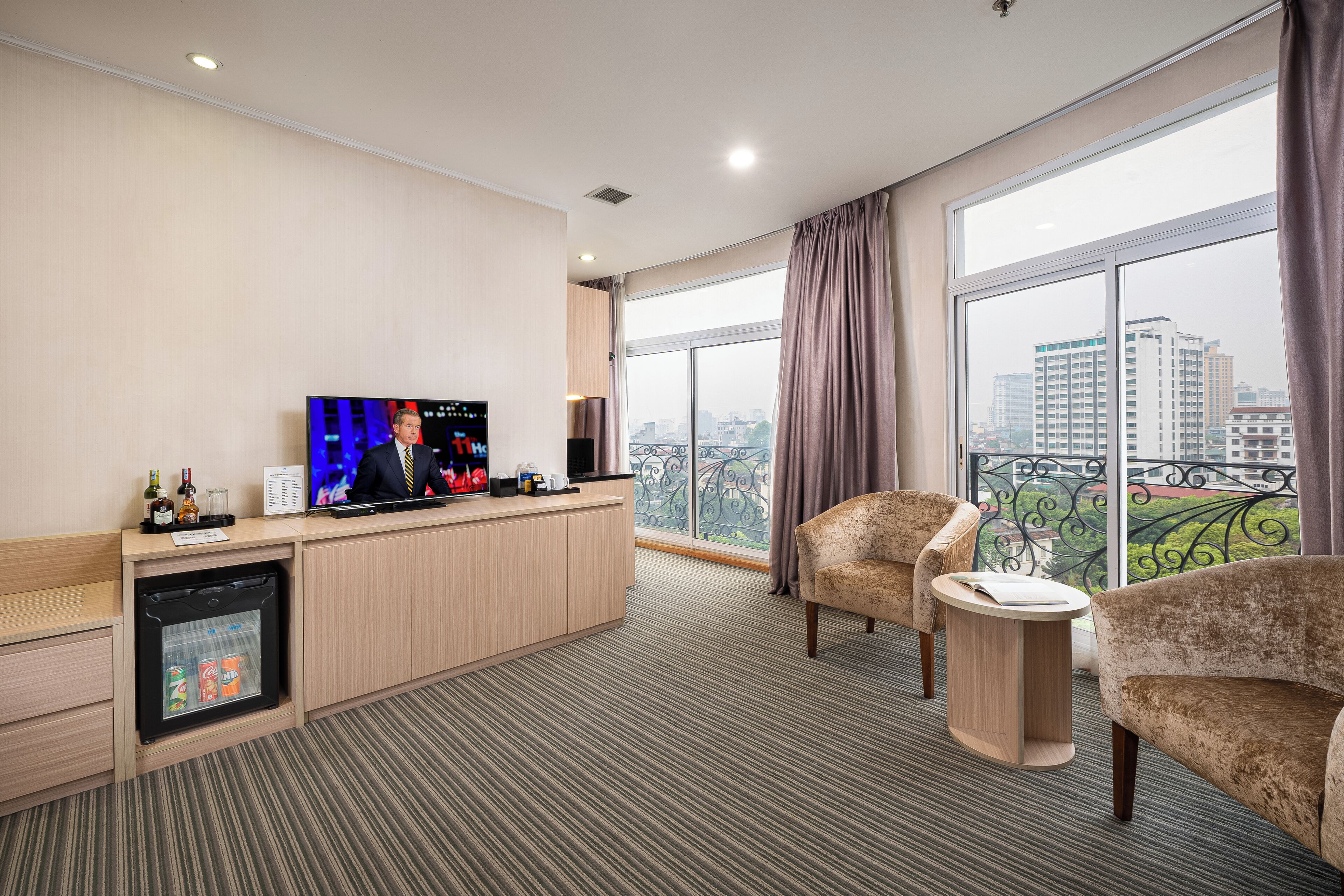 A hotel room with a TV, minibar, seating area, and a city view.