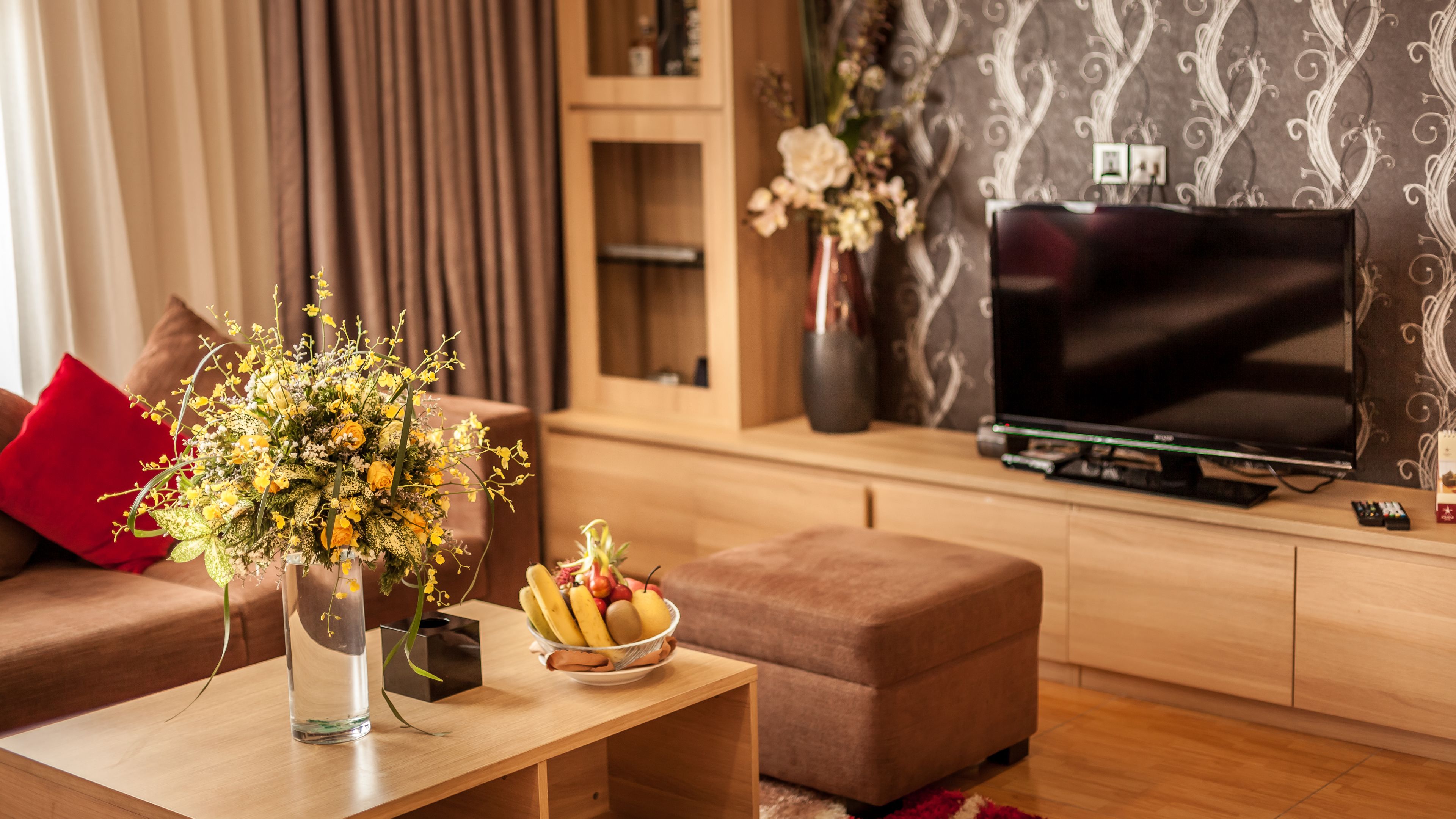 A modern living room with a sofa, a coffee table with a vase of flowers, a fruit bowl, and a flat-screen TV on a wooden cabinet.