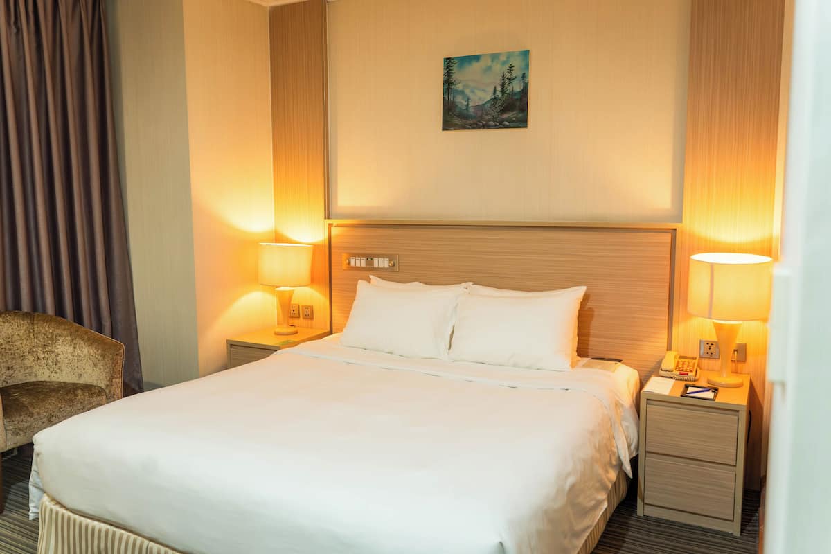 Hotellihuone, jossa on suuri sänky, kaksi yöpöytää valaisimineen, tuoli ja taulu seinällä.