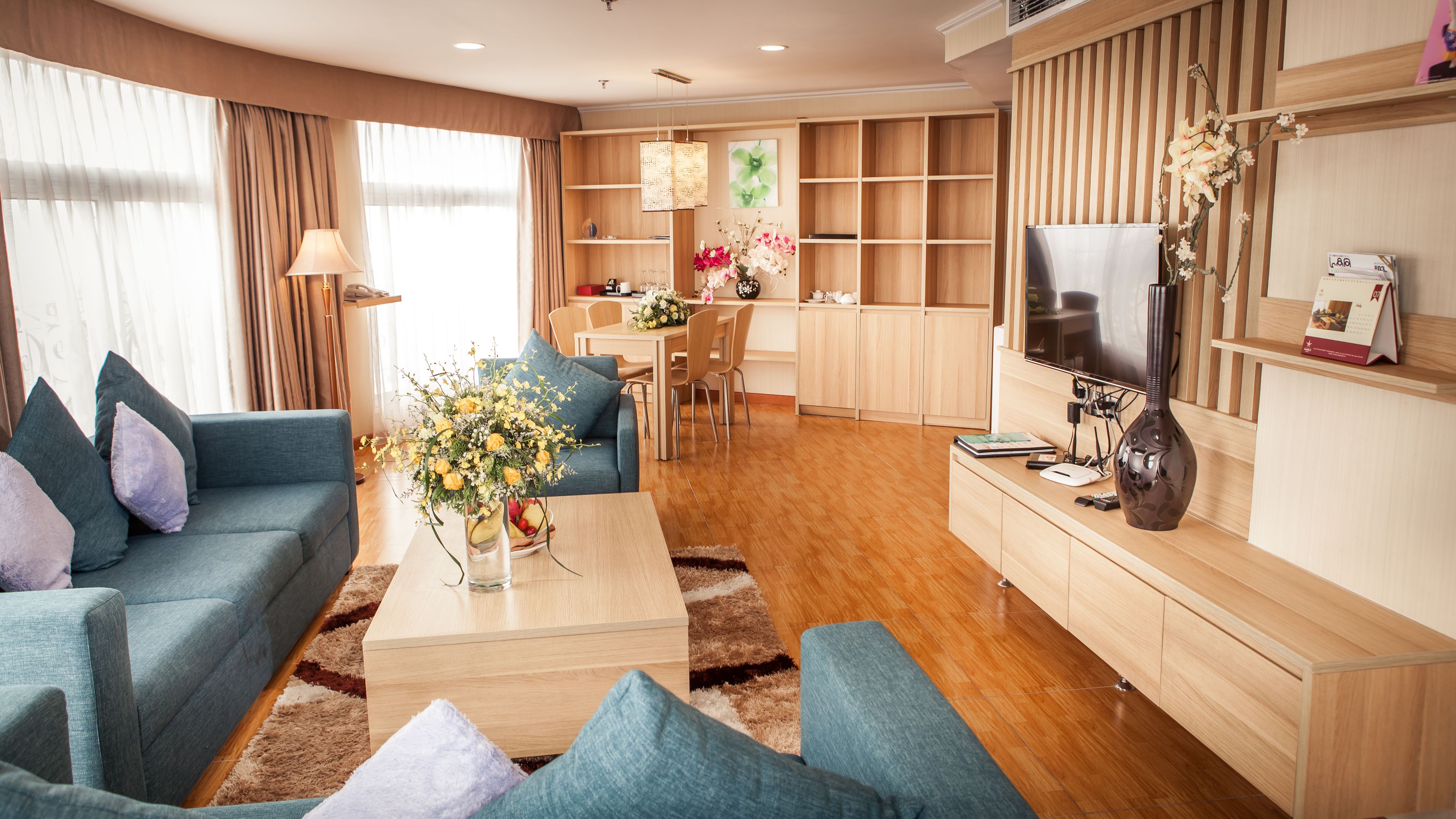 A modern living room with a sofa, coffee table, and TV. There is a dining area with chairs and a table, and a kitchen area with cabinets and a TV.