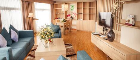 A modern living room with a sofa, coffee table, and TV. There is a dining area with chairs and a table, and a kitchen area with cabinets and a TV.