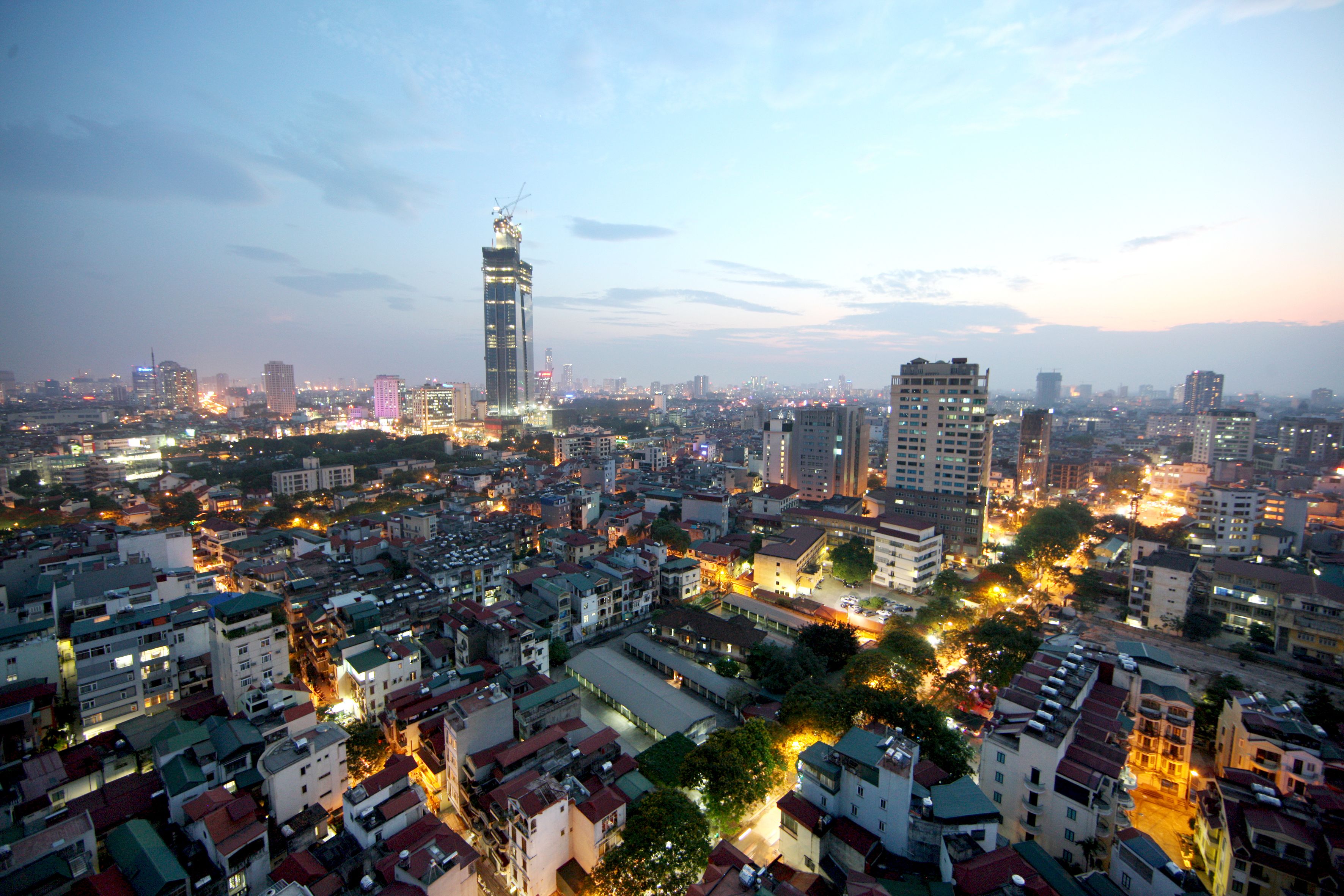 A cityscape at dusk with a prominent skyscraper under construction, surrounded by densely packed buildings and illuminated streets.