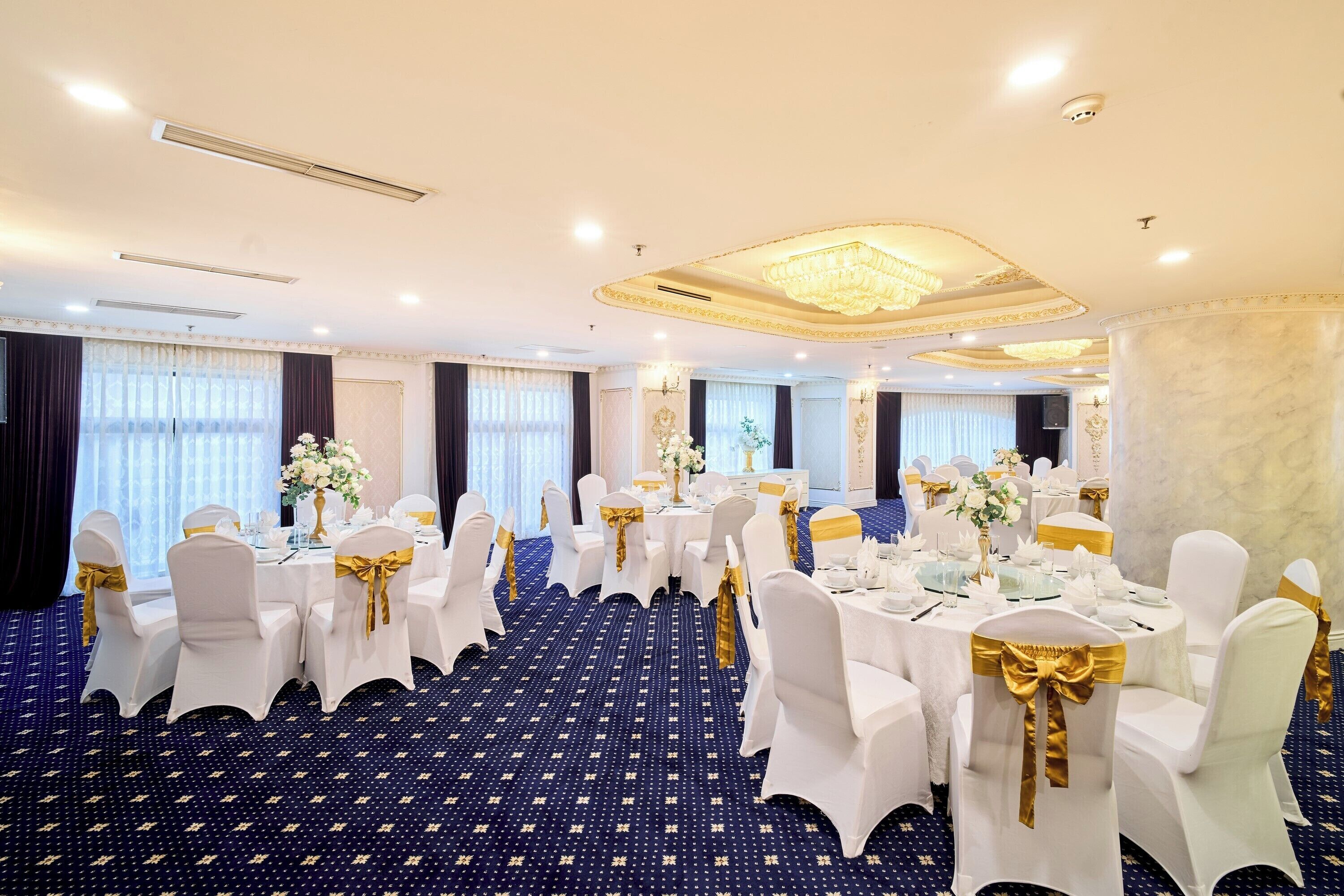 A banquet hall with round tables, white chairs with gold sashes, and a blue carpet with a star pattern.