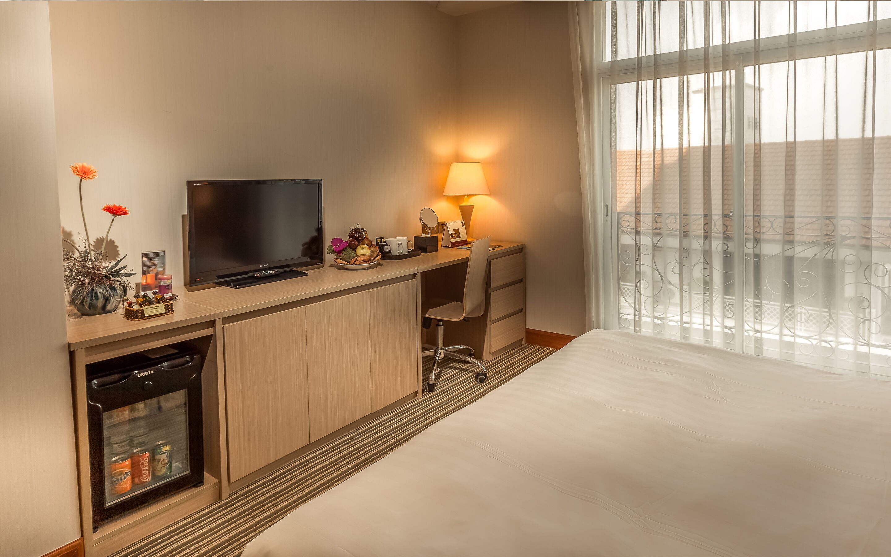 Habitación de hotel con cama, escritorio con televisor, lámpara, minibar y ventana con persianas.