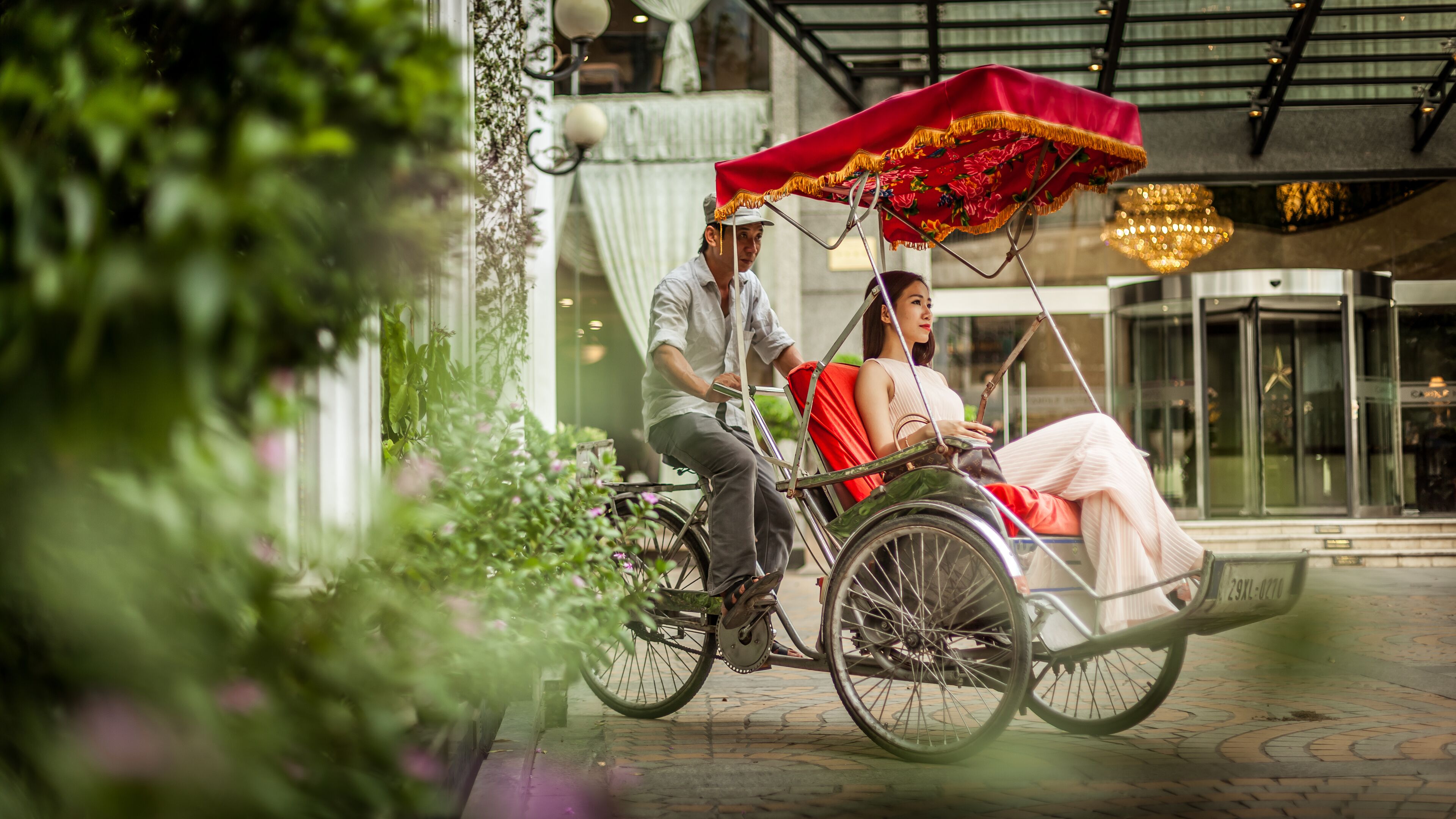 A couple in a rickshaw with a red canopy, parked in front of a building with a glass entrance and a star decoration above the door.