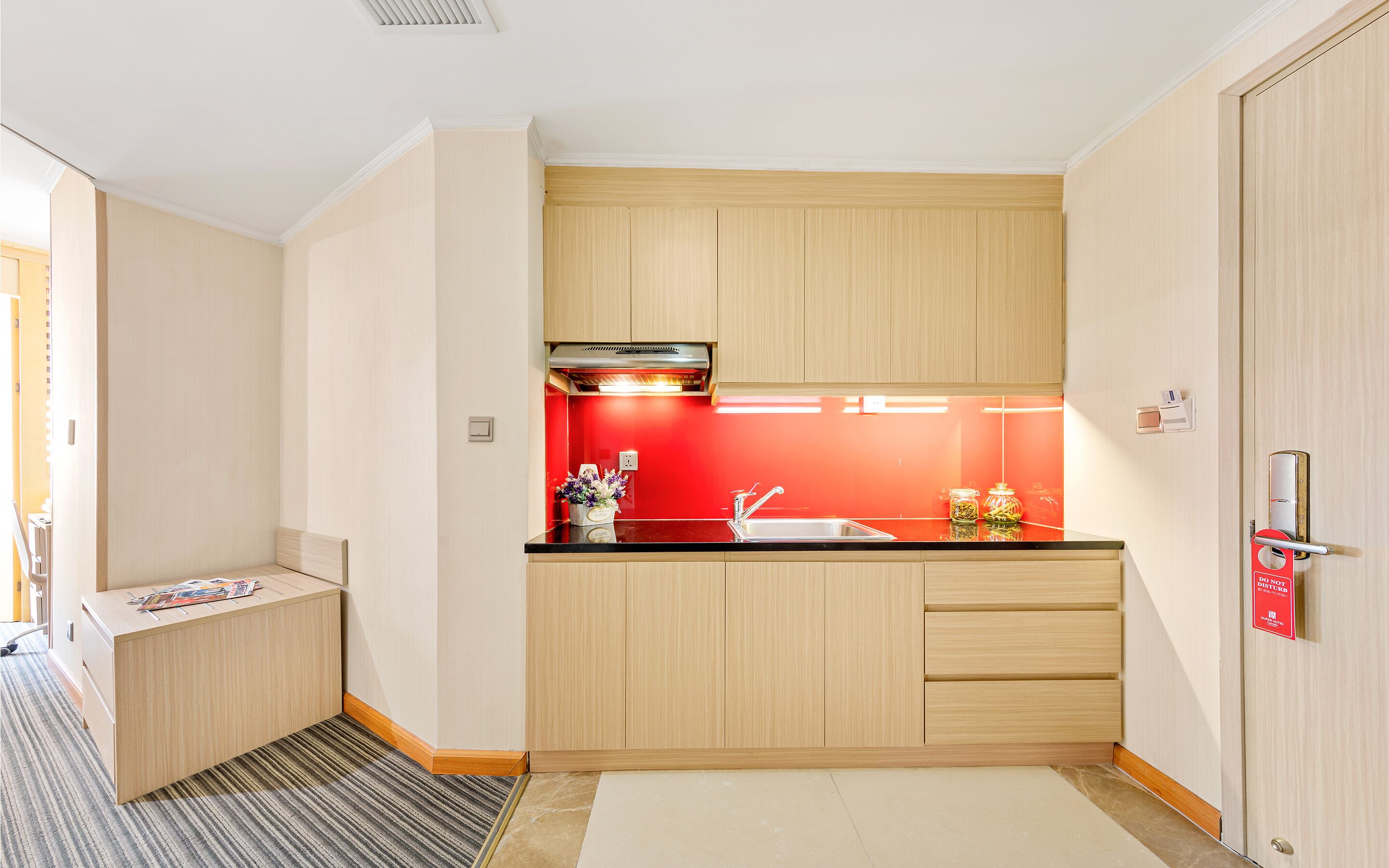 Cocina compacta con armarios de madera, encimera negra, salpicadero rojo y horno empotrado.
