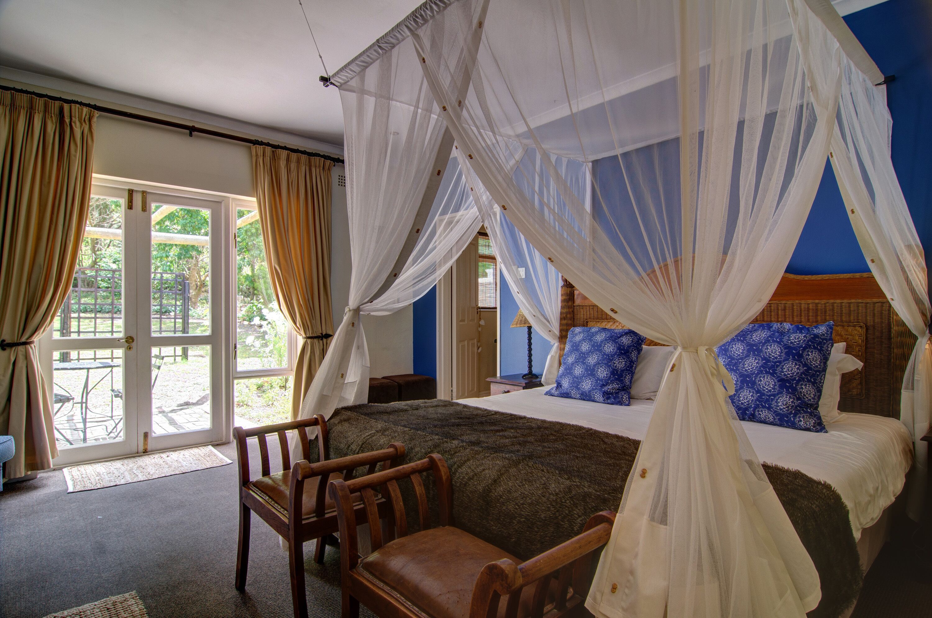 A bedroom with a four-poster bed, a wooden chair, and a view through a glass door to an outdoor area.