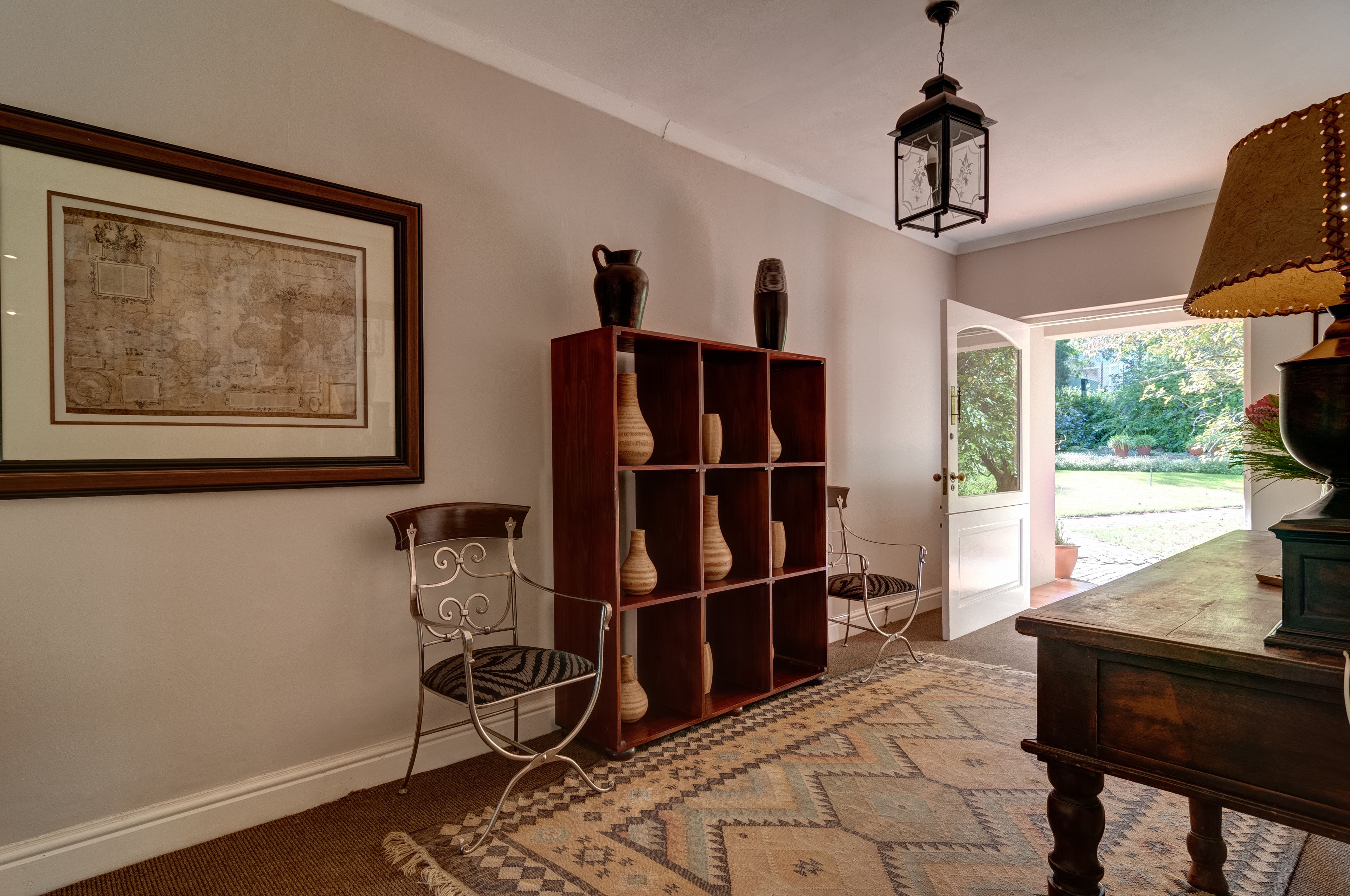 A hallway with a patterned rug, a wooden bookshelf with vases, a metal chair, and a framed map on the wall.