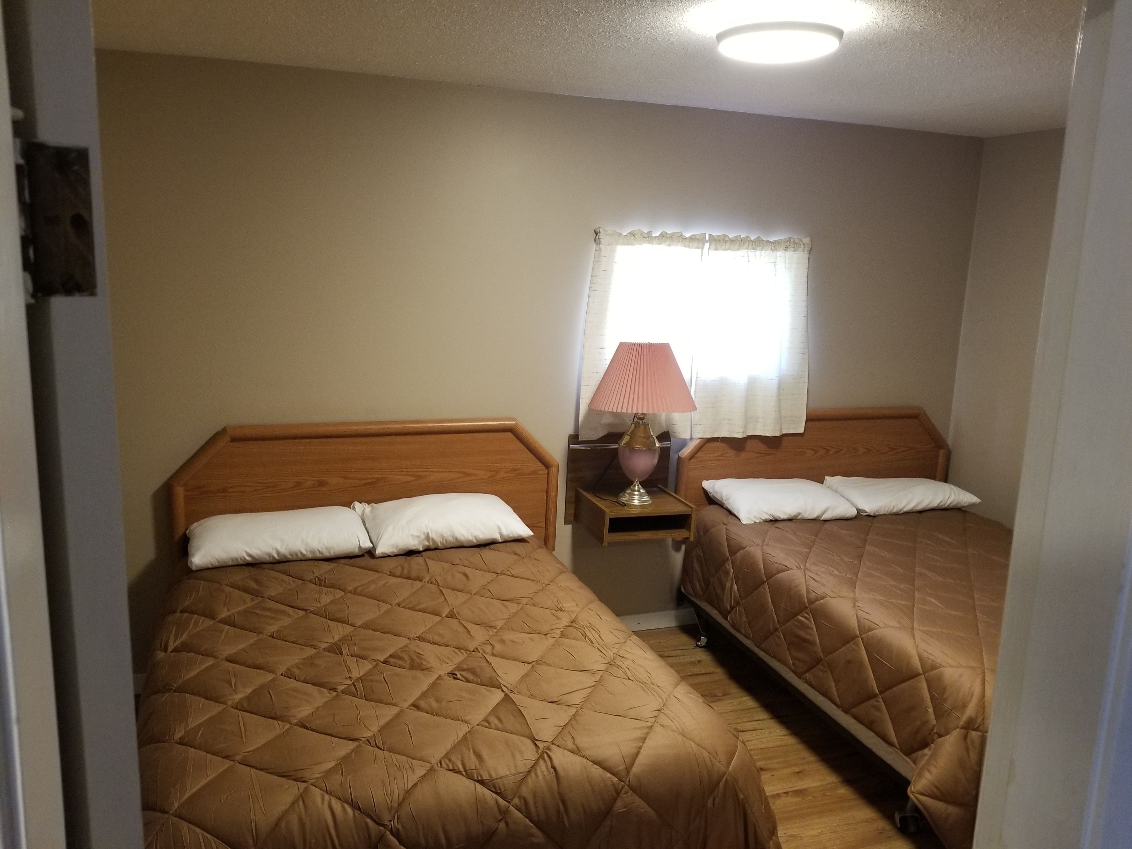 A room with two beds, each with a wooden headboard and a quilted brown bedspread. There is a nightstand with a lamp between the beds.