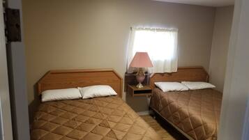 A room with two beds, each with a wooden headboard and a quilted brown bedspread. There is a nightstand with a lamp between the beds.