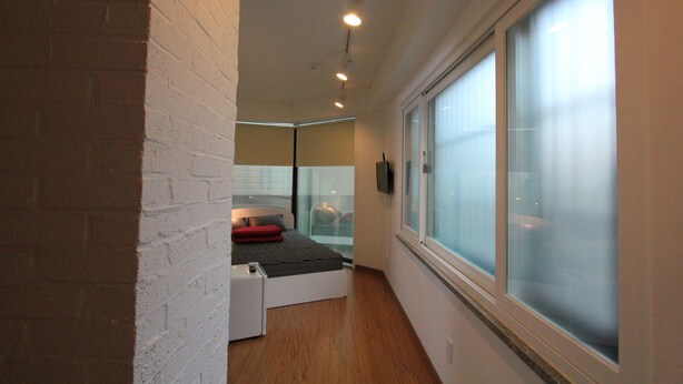 A narrow hallway with a bed at the end, a large window, and a white brick wall. - B my guesthouse - Hostel (Seoul)