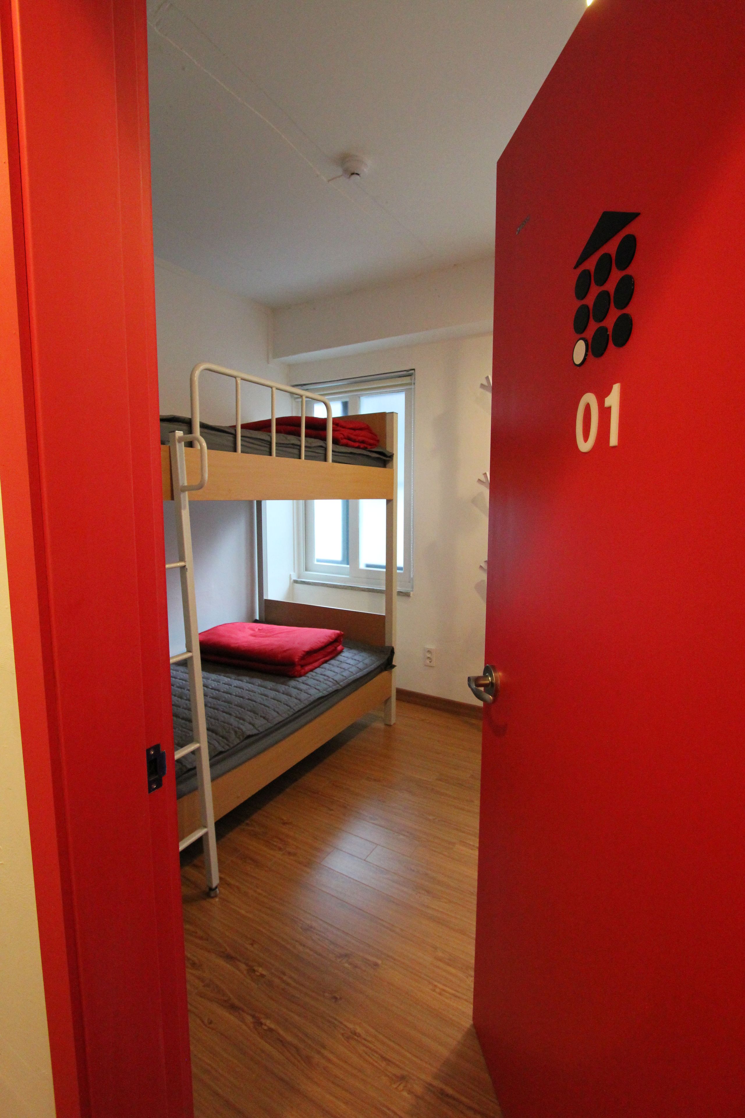 A bunk bed room with a red door labeled "01" and a window with a view of the outside.