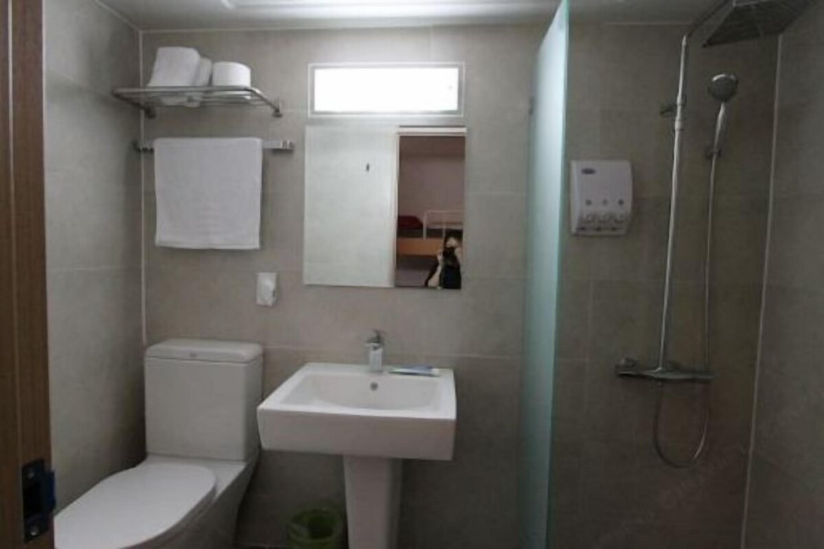 A small bathroom with a toilet, sink, and shower. There is a mirror above the sink and a shelf with towels.