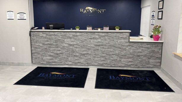 A reception area with a dark blue wall, a stone-textured counter, and a logo that reads "Baymont by Wyndham". - Baymont by Wyndham Dickinson (Dickinson)