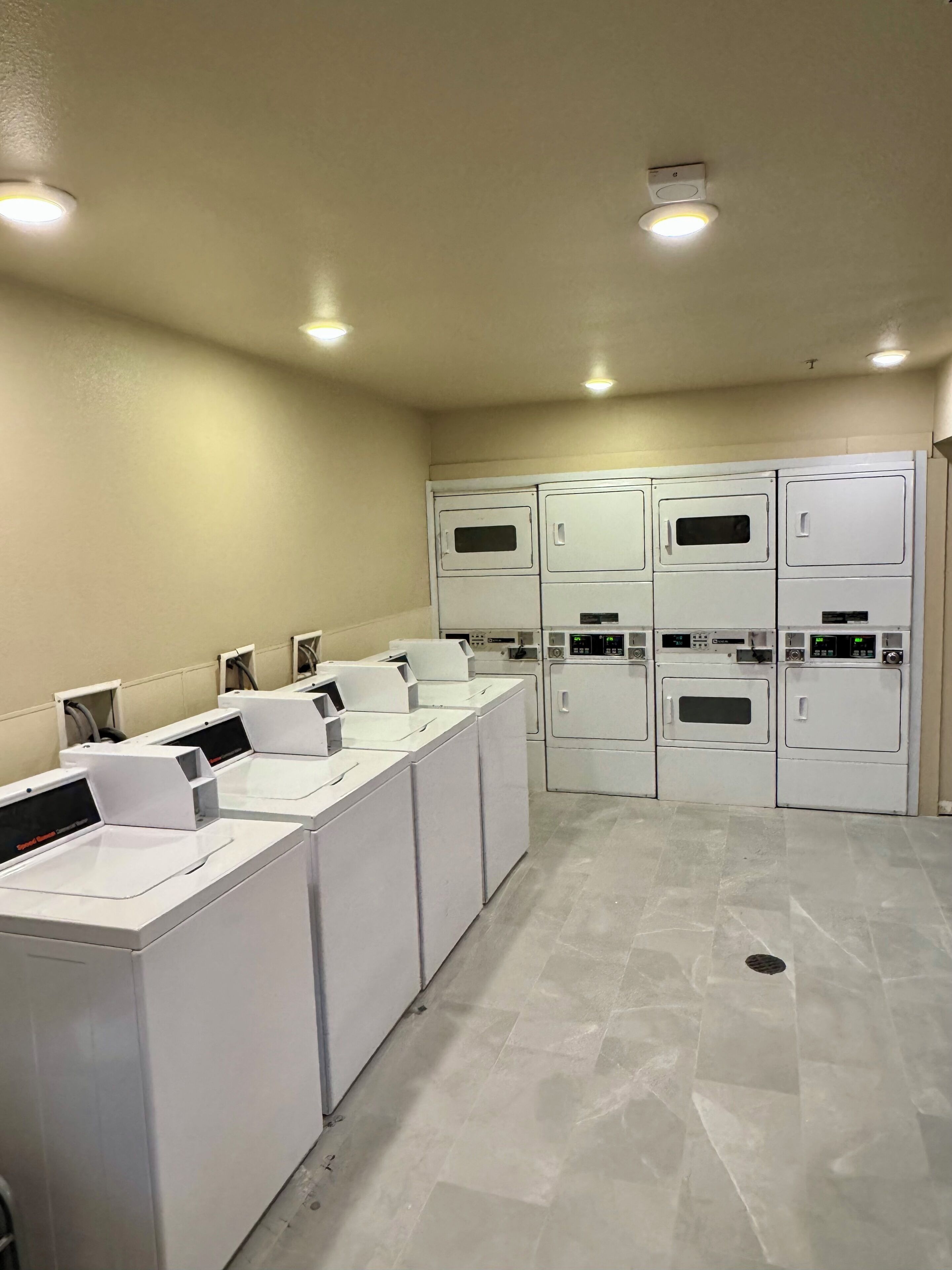 A laundry facility with multiple washing machines and dryers, a tiled floor, and recessed ceiling lights.