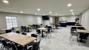 A spacious dining area with multiple tables and chairs, a kitchen area with a counter and appliances, and large windows providing natural light. - Baymont by Wyndham Dickinson (Dickinson)