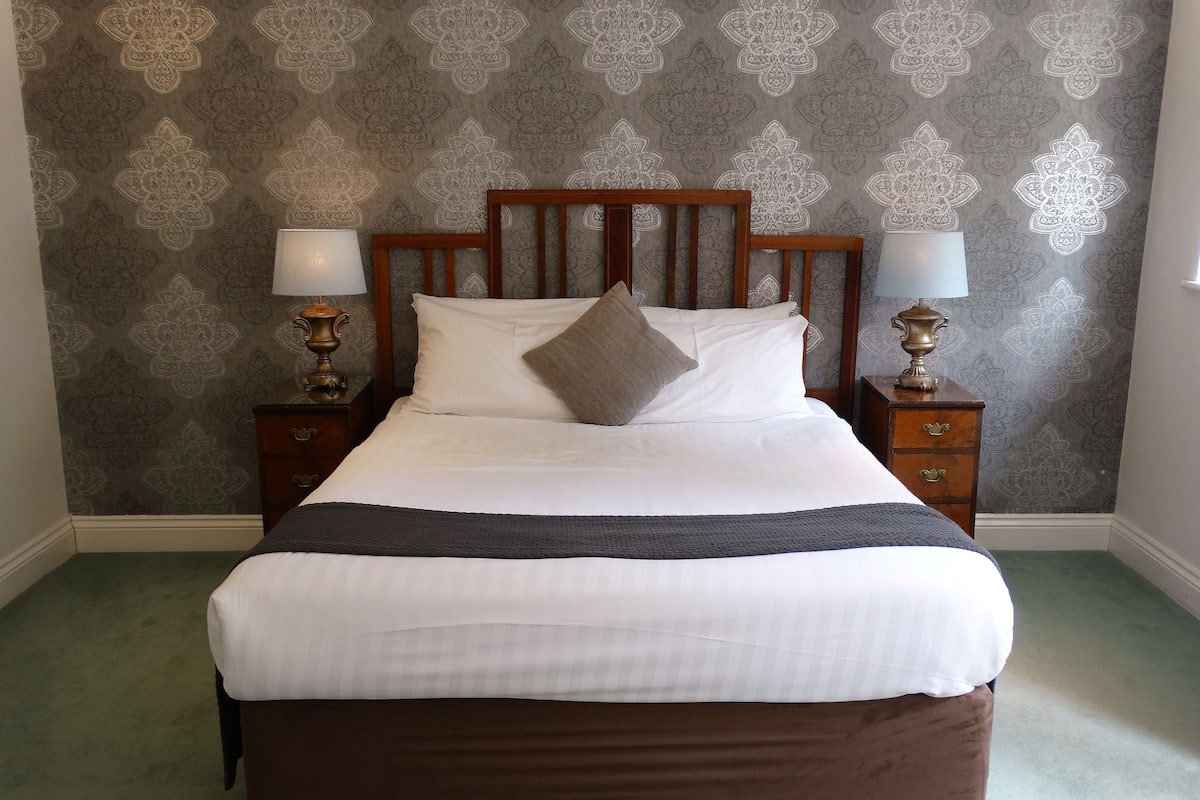 Un lit bien fait, avec une tête de lit en bois, du linge de lit blanc et un plaid marron. Deux tables de chevet, ornées de lampes et de vases décoratifs, se trouvent de part et d’autre du lit. Le mur est recouvert d’un papier peint à motifs.