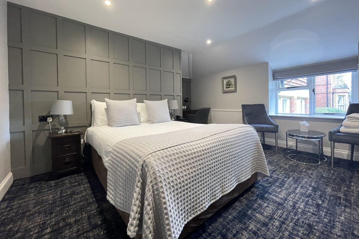 Une chambre d’hôtel avec un grand lit, une table de chevet avec une lampe, une chaise, une petite table avec une boîte de mouchoirs et une fenêtre donnant sur des immeubles.