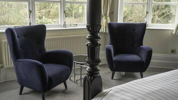 A bedroom with two dark blue armchairs, a tall black column, a bed with a striped bedspread, and a window with a view of trees.