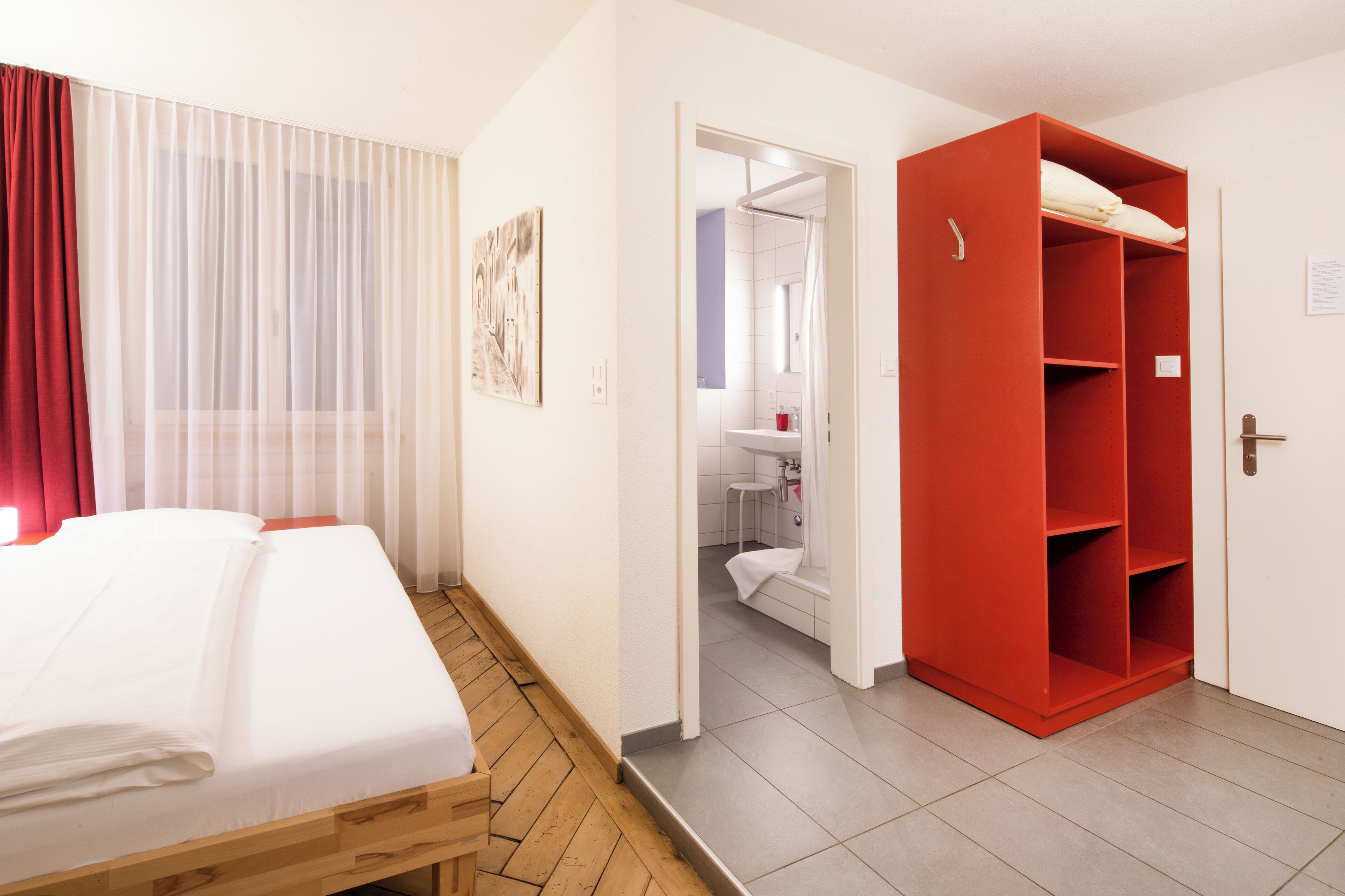 Une chambre d’hôtel avec un lit, une armoire rouge, une salle de bain avec douche et un tableau au mur.
