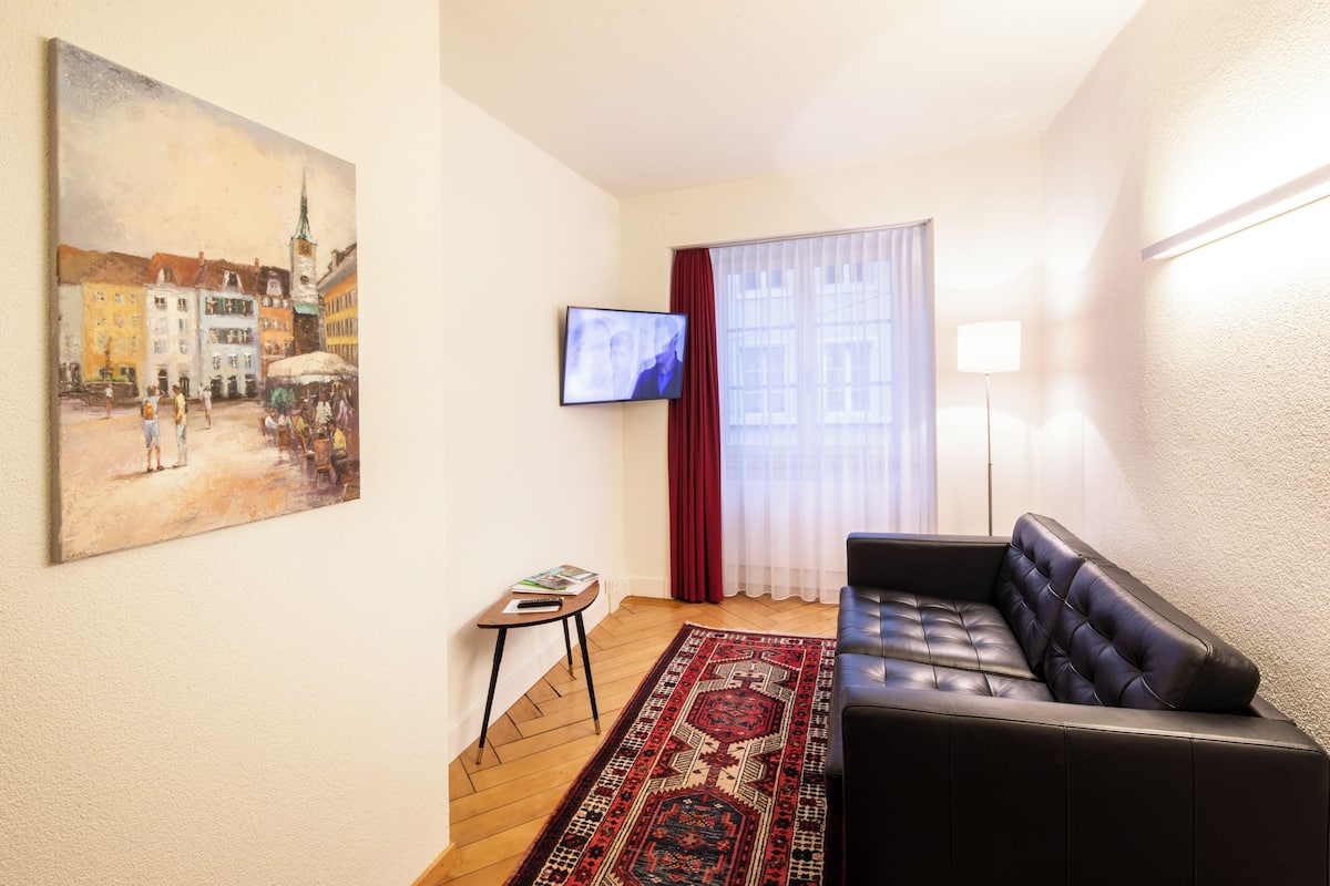 A living room with a black leather sofa, a patterned rug, a painting on the wall, a television, and a window with curtains.