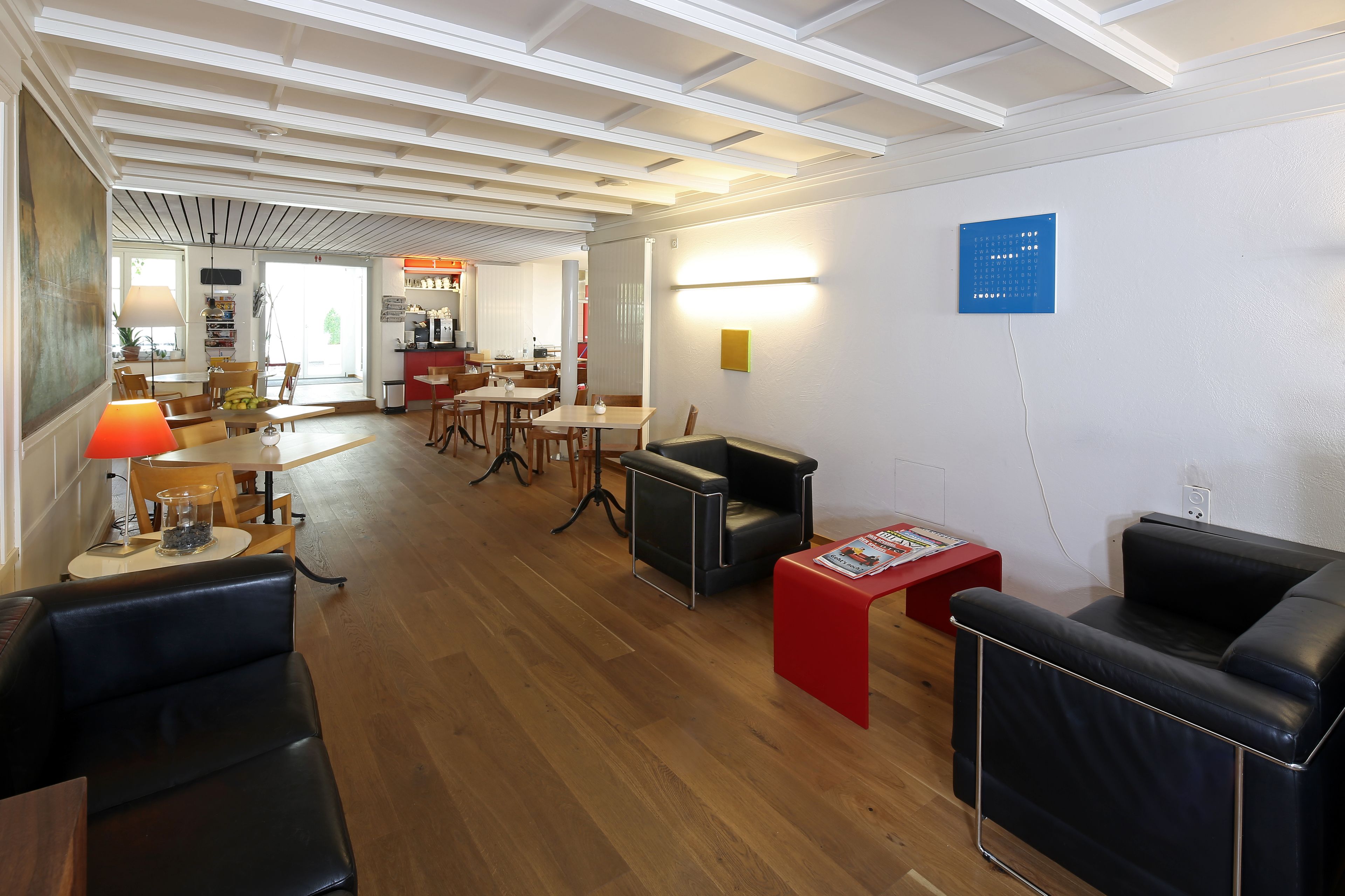 Een moderne hotellobby met houten vloeren, zwarte lederen zitplaatsen, een rode accenttafel en een licht plafond met zichtbare balken.