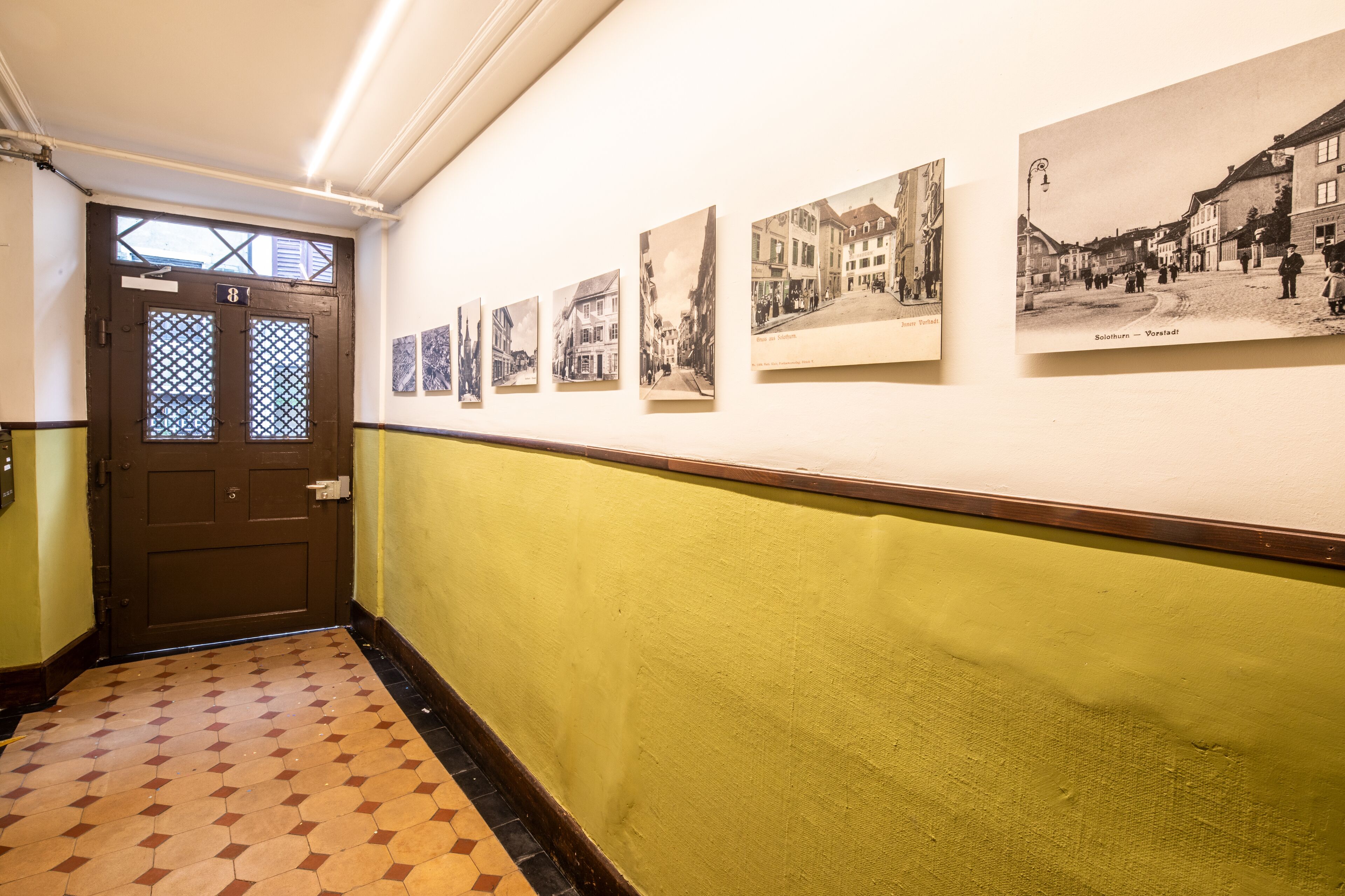 A hallway with a patterned floor, a door with a number, and several black and white photographs on the wall.