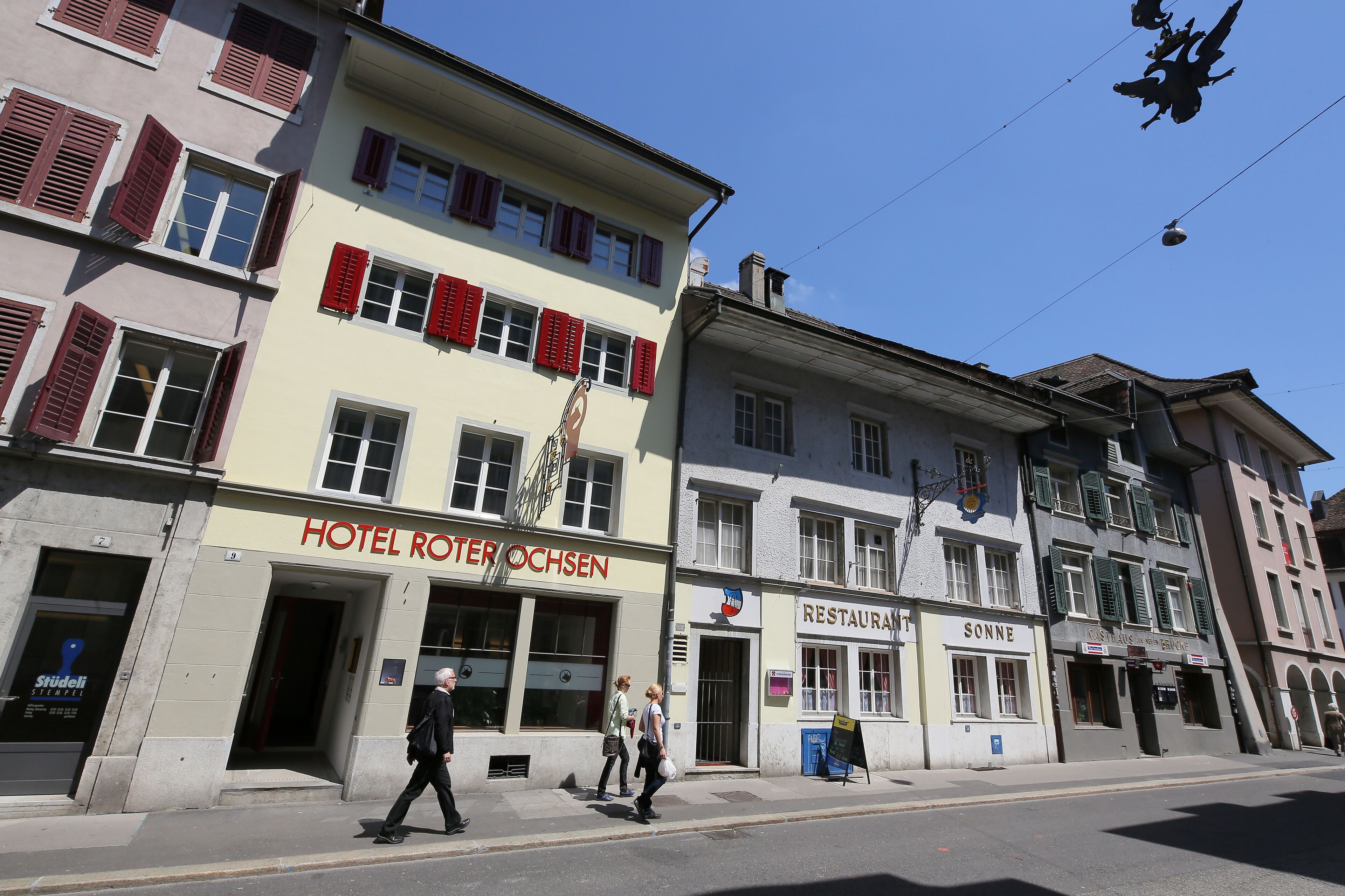 A hotel named 'HOTEL RÖTHERÖCHSEN' with a traditional facade, a restaurant 'RESTAURANT SONNE' across the street, and a 'Café' with outdoor seating.