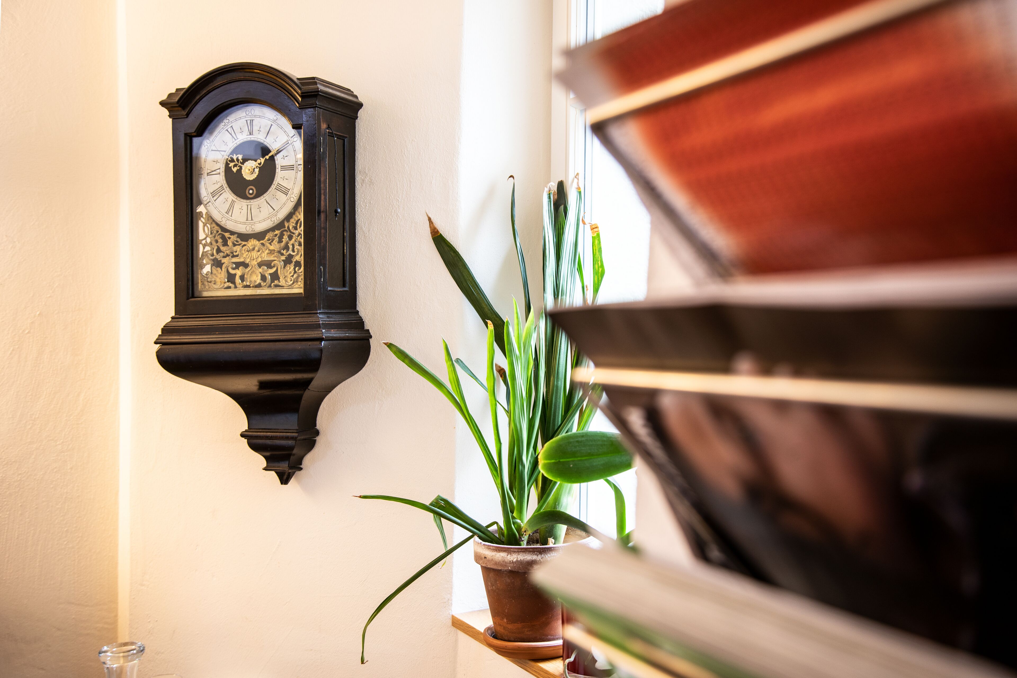 Un reloj de pie adosado a la pared, una planta en maceta y un piano.