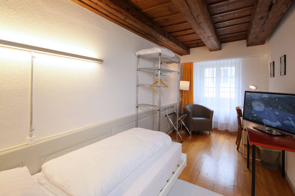 A hotel room with a bed, a desk with a computer, a chair, a shelf with clothes, and a window with curtains.