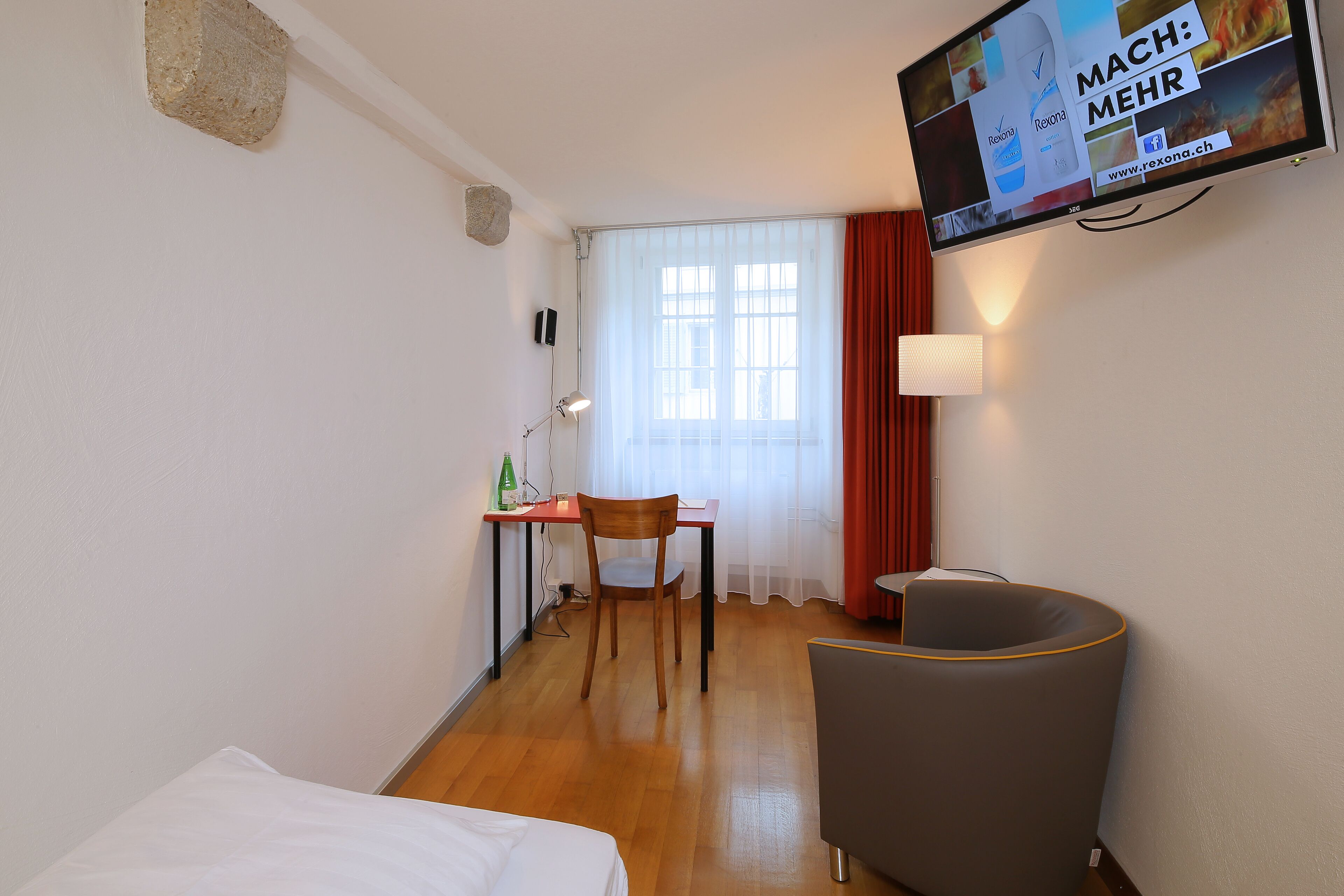 A hotel room with a bed, a desk, a chair, a TV mounted on the wall, and a window with curtains.
