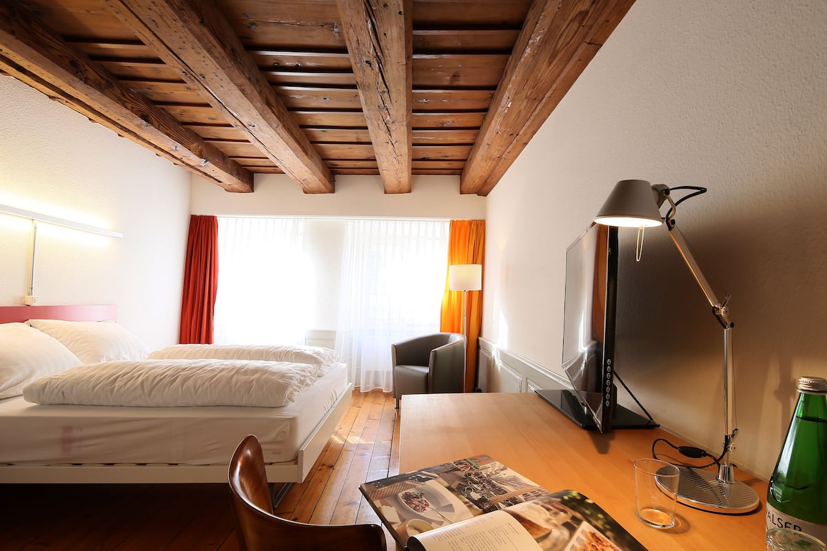 A hotel room with a wooden ceiling, a bed with white linens, a desk with a lamp, a chair, a computer, and a bottle of water.