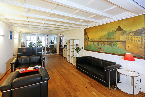 Un vestíbulo de hotel con piso de madera, sofás de cuero negro, una mesa roja y un gran mural de un castillo.