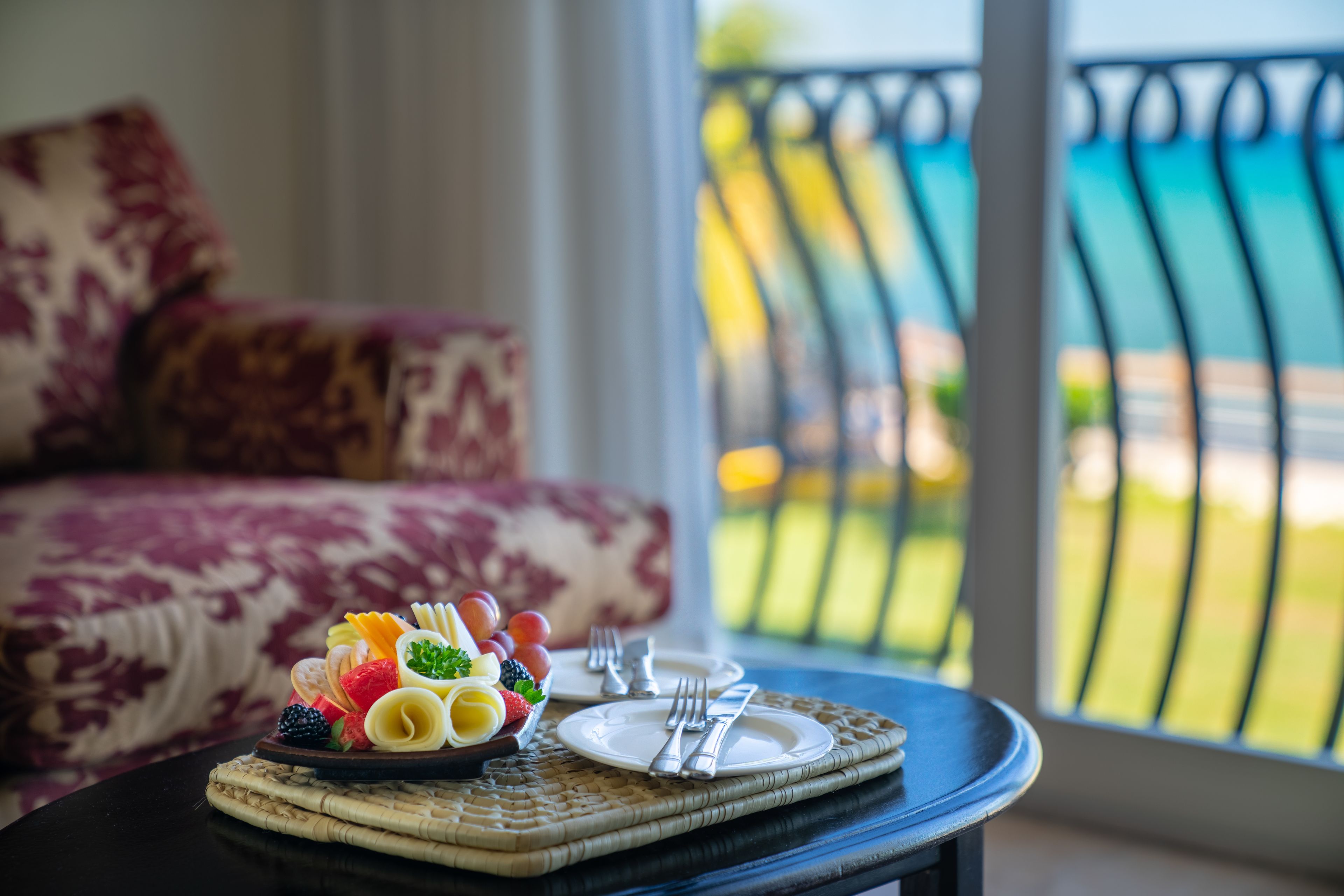 A dining table with a fruit platter and cutlery, a sofa with a patterned cushion, and a view of a balcony with a railing.