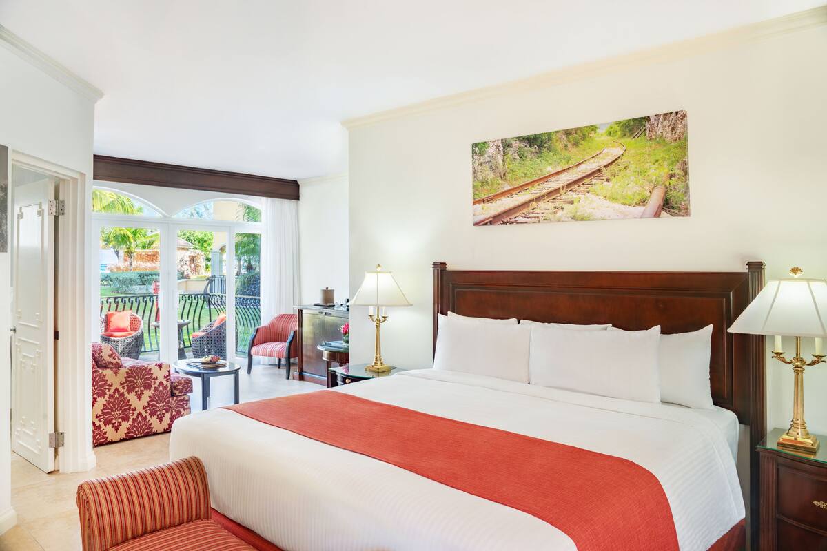Une chambre d’hôtel avec un grand lit, un tableau représentant une voie ferrée, un balcon avec des meubles d’extérieur et une vue sur la verdure.