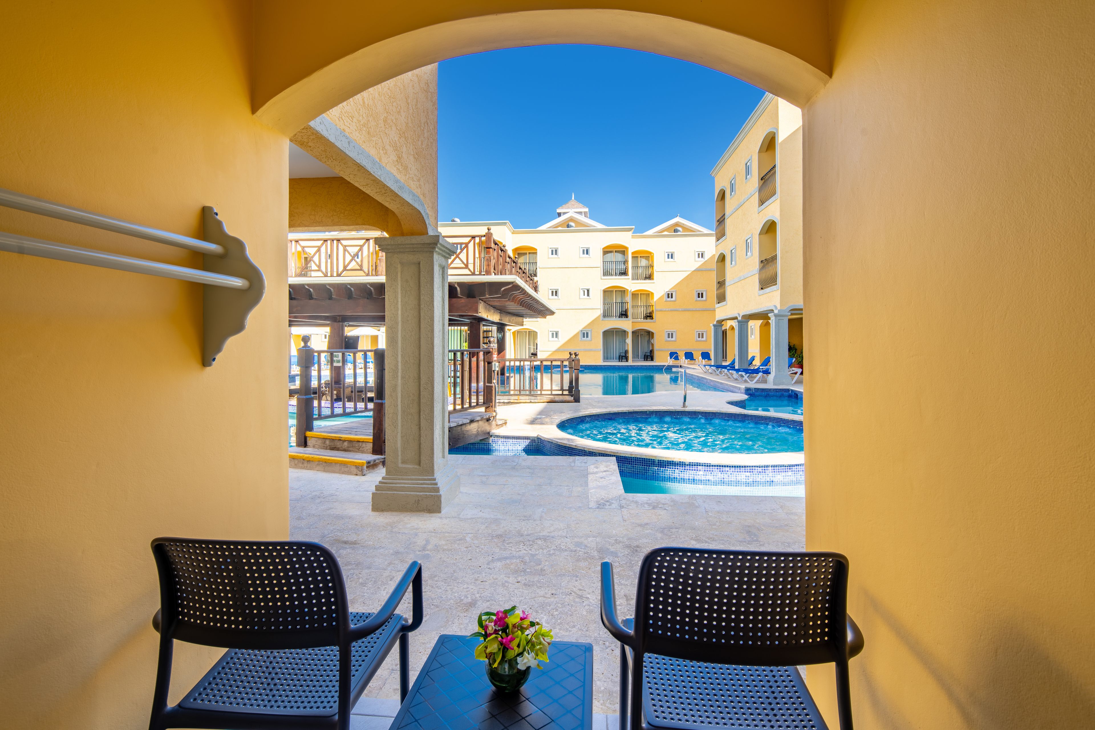 A view through an archway of a pool area with two chairs and a table, a pool, and buildings in the background.